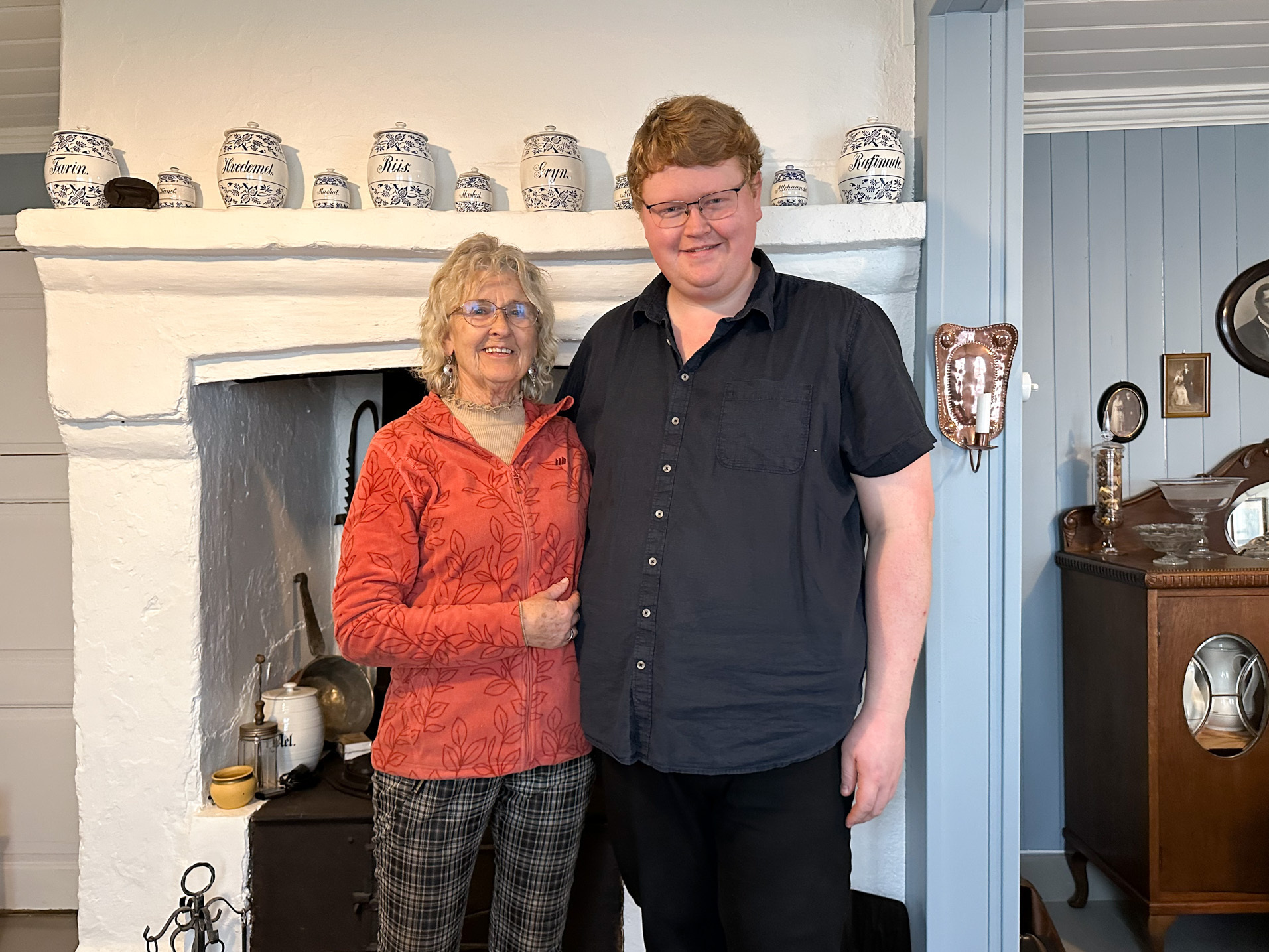 Kvinne i 70-årene står sammen med en høy mann i 30-årene inne på et kjøkken i gammel stil.