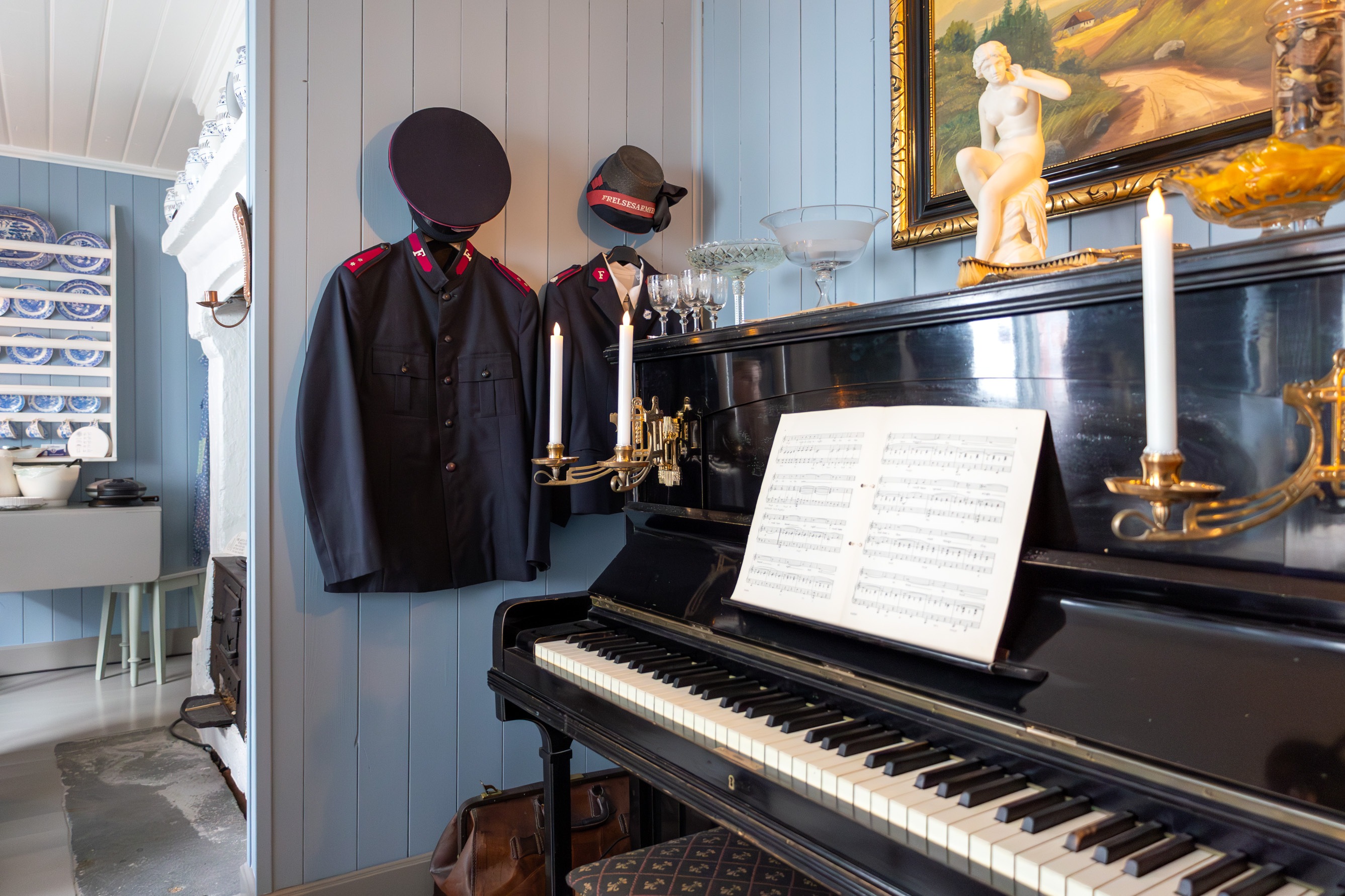 Gammelt piano med lysestake montert på hver side over tangentene. Over henger et maleri med tykk gullramme og i kroken heger et par uniformer.