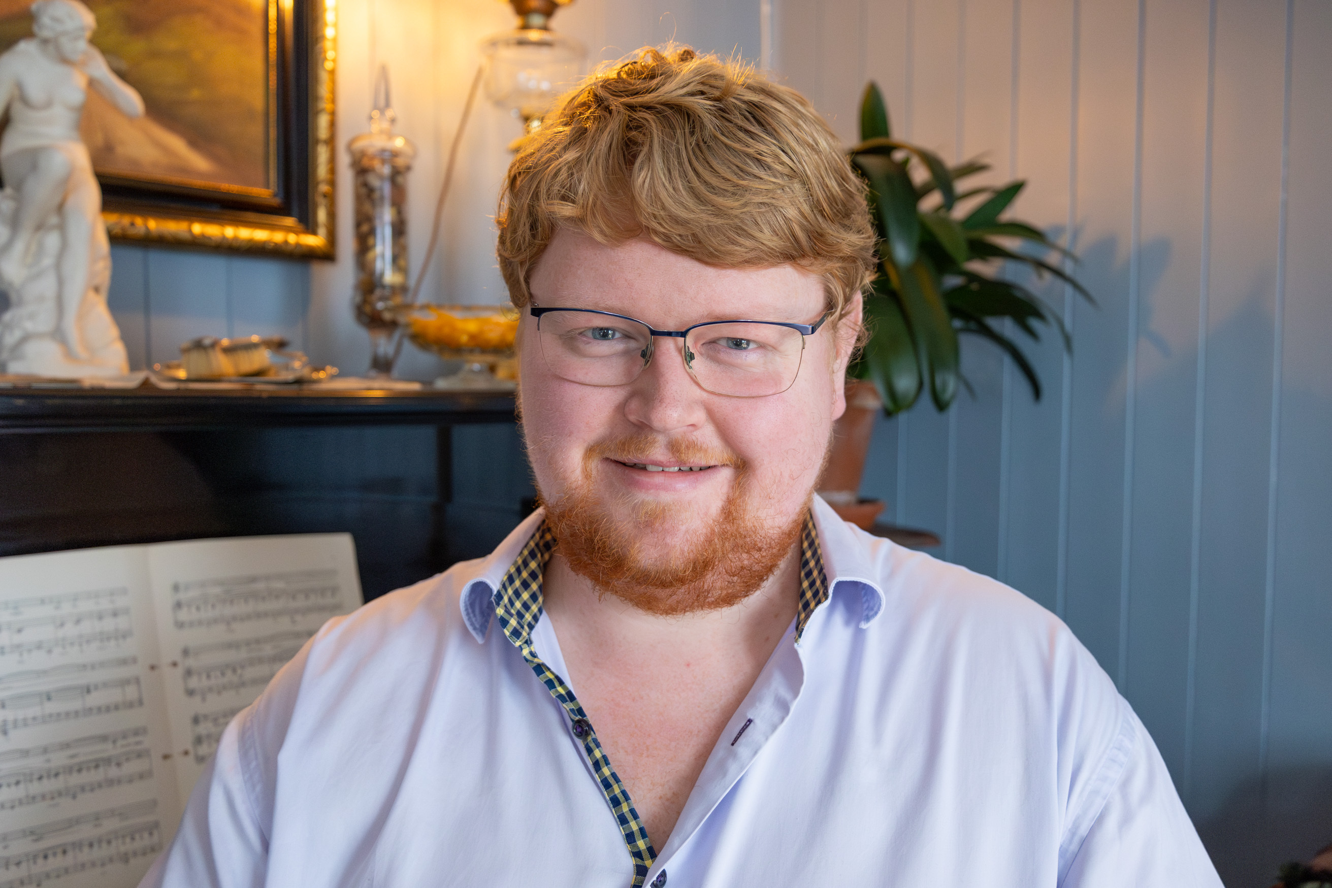 Ung mann med briller og skjegg smiler inn i kamera. Han sitter ved pianoet og har på seg penskjorte.