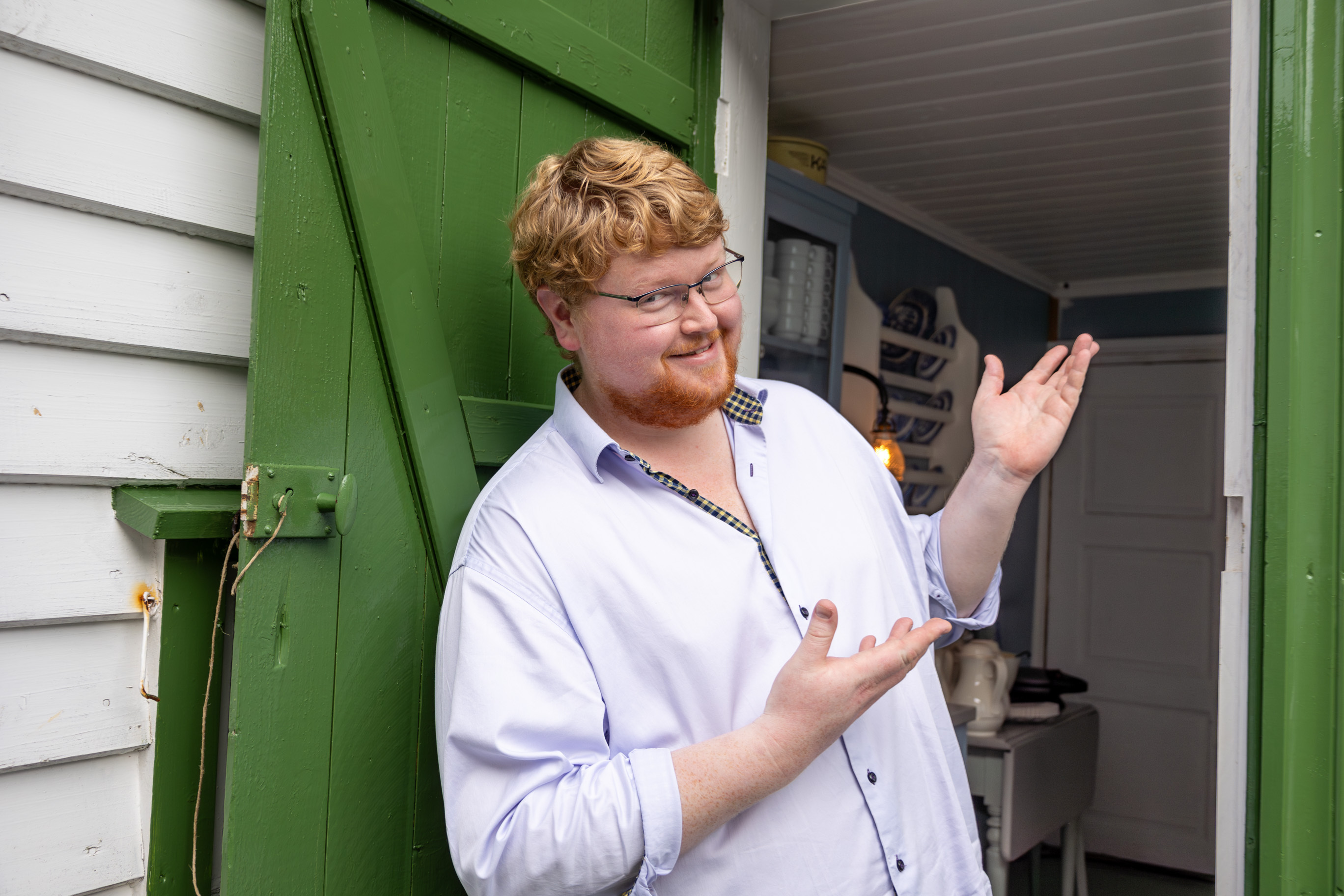 Yngre mann med briller, hår og skjegg holder opp hendene og viser velkommen inn i et rom. Han står i døråpningen til et gammelt hus.