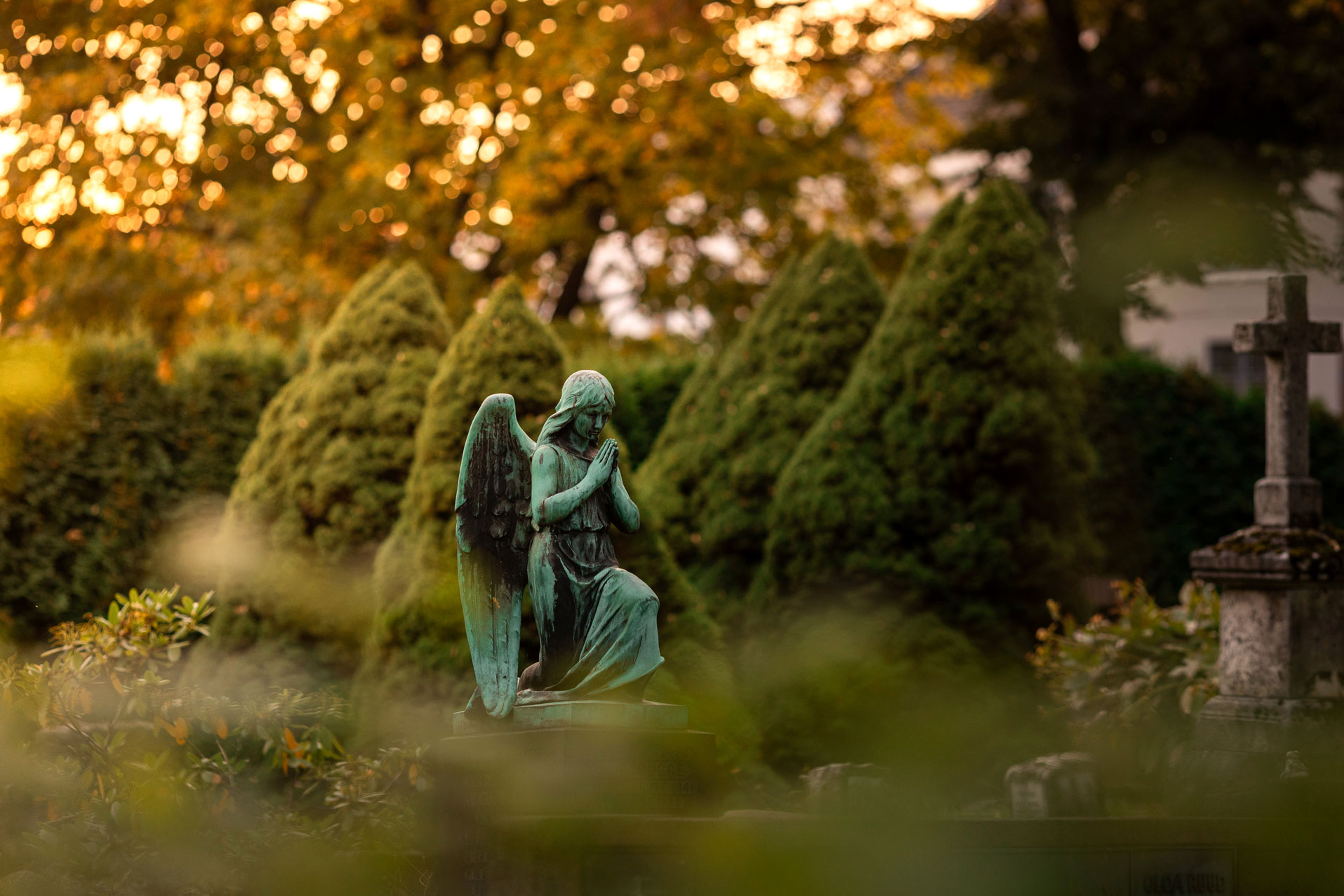 På en gravplass i høstfarger er fokus stilt inn på en statue av en grønn engel i bronse som kneler og ber.