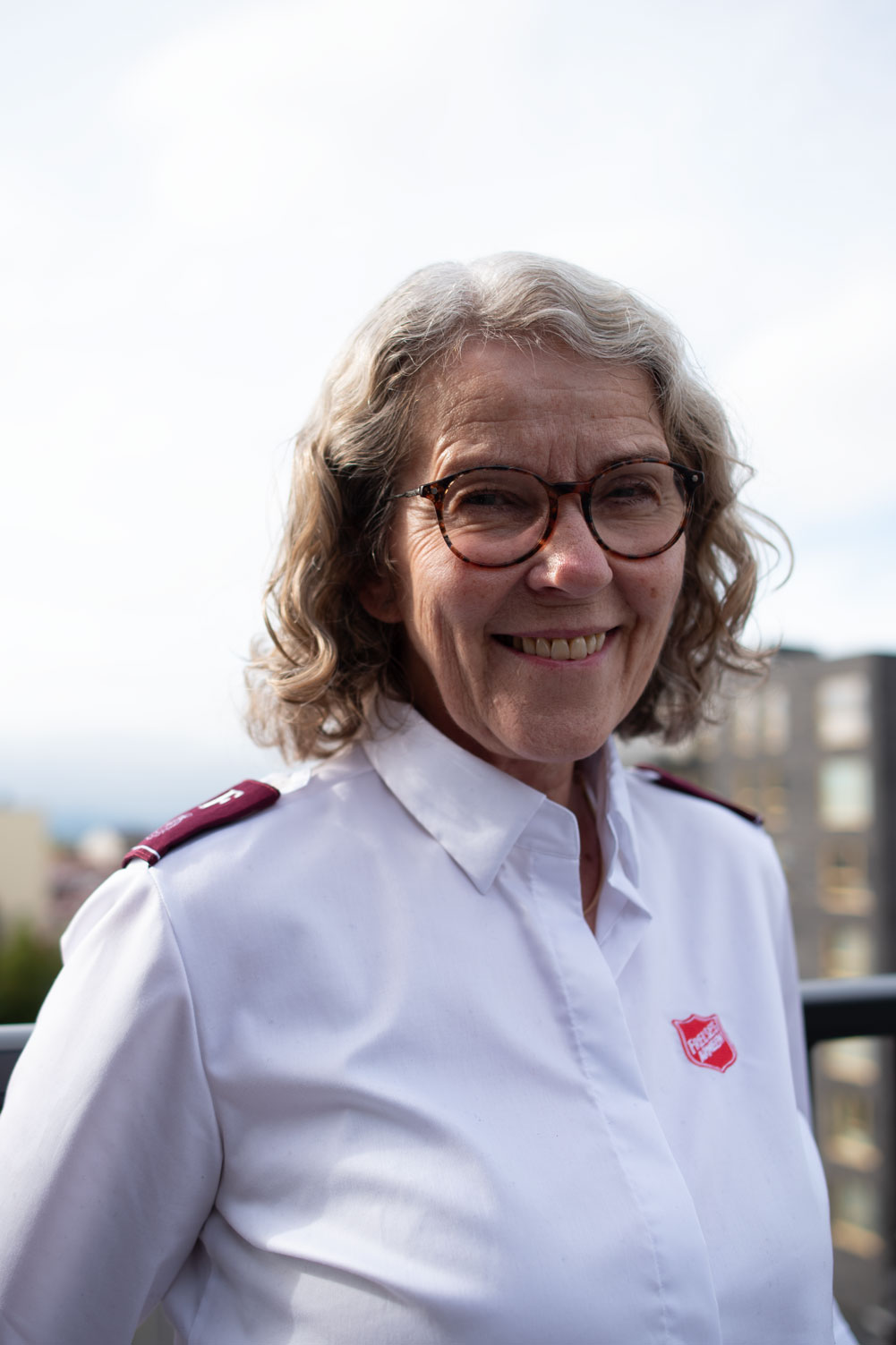 Kvinne med grå krøller og briller står i hvit uniformsskjorte med rød logo fra Frelsesarmeen. I bakgrunnen sees grå blokker av nyere dato.