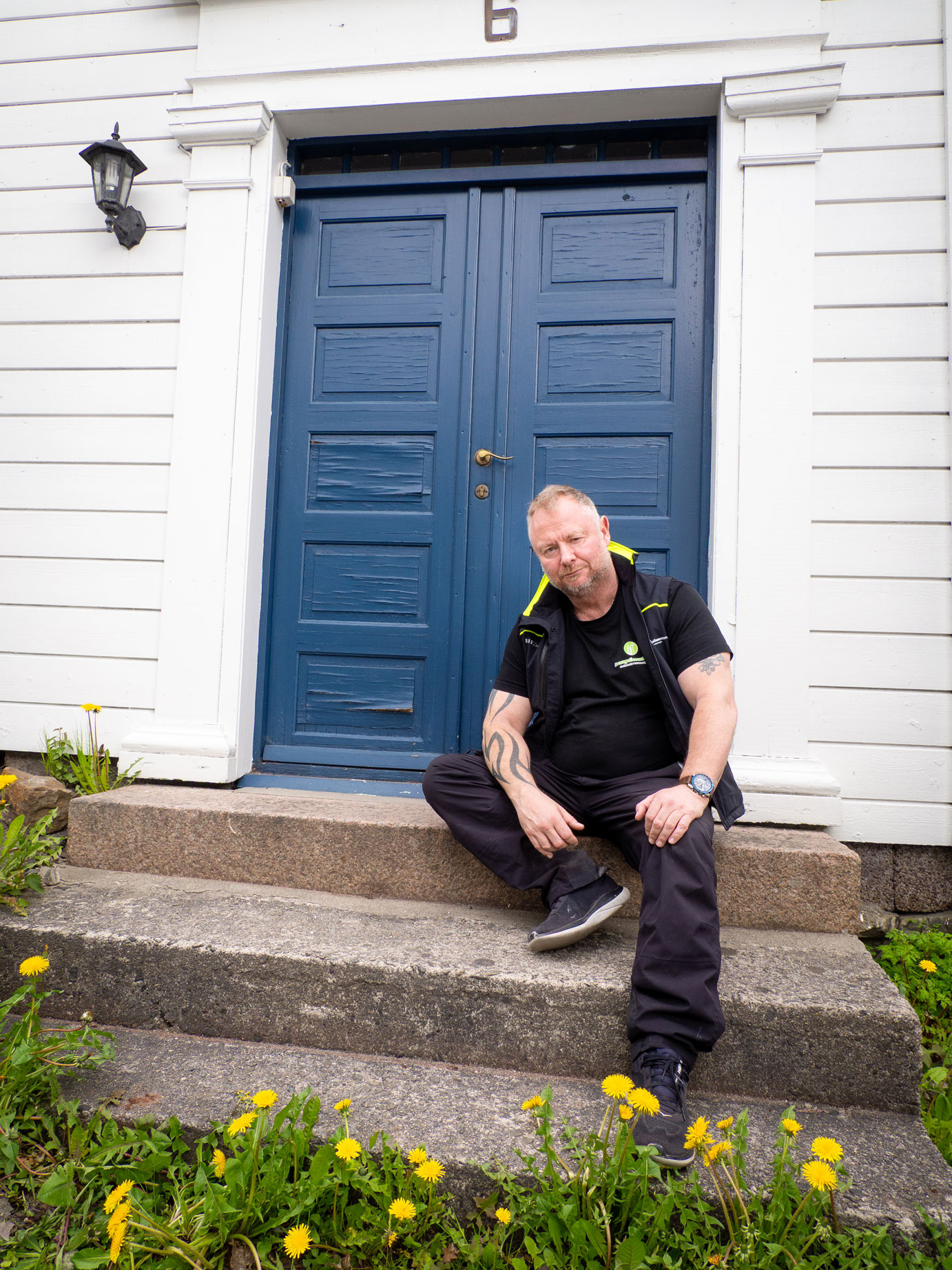 Foto: Mann med tatoveringer sitter på en steintrapp opp til et gammelt hvitt hus med blå dør.