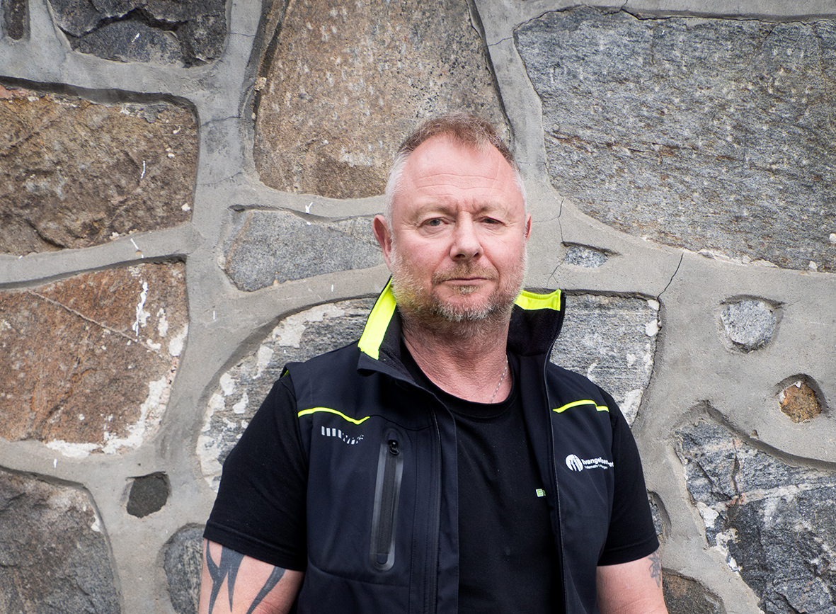 Foto: Mann i 50-årene med tatoveringer på begge armene står mot en gammel mur av steiner. Han har grått, kortklippet hår og todagers skjegg. Han har på seg en mørk T-skjorte og en vest med gule detaljer.