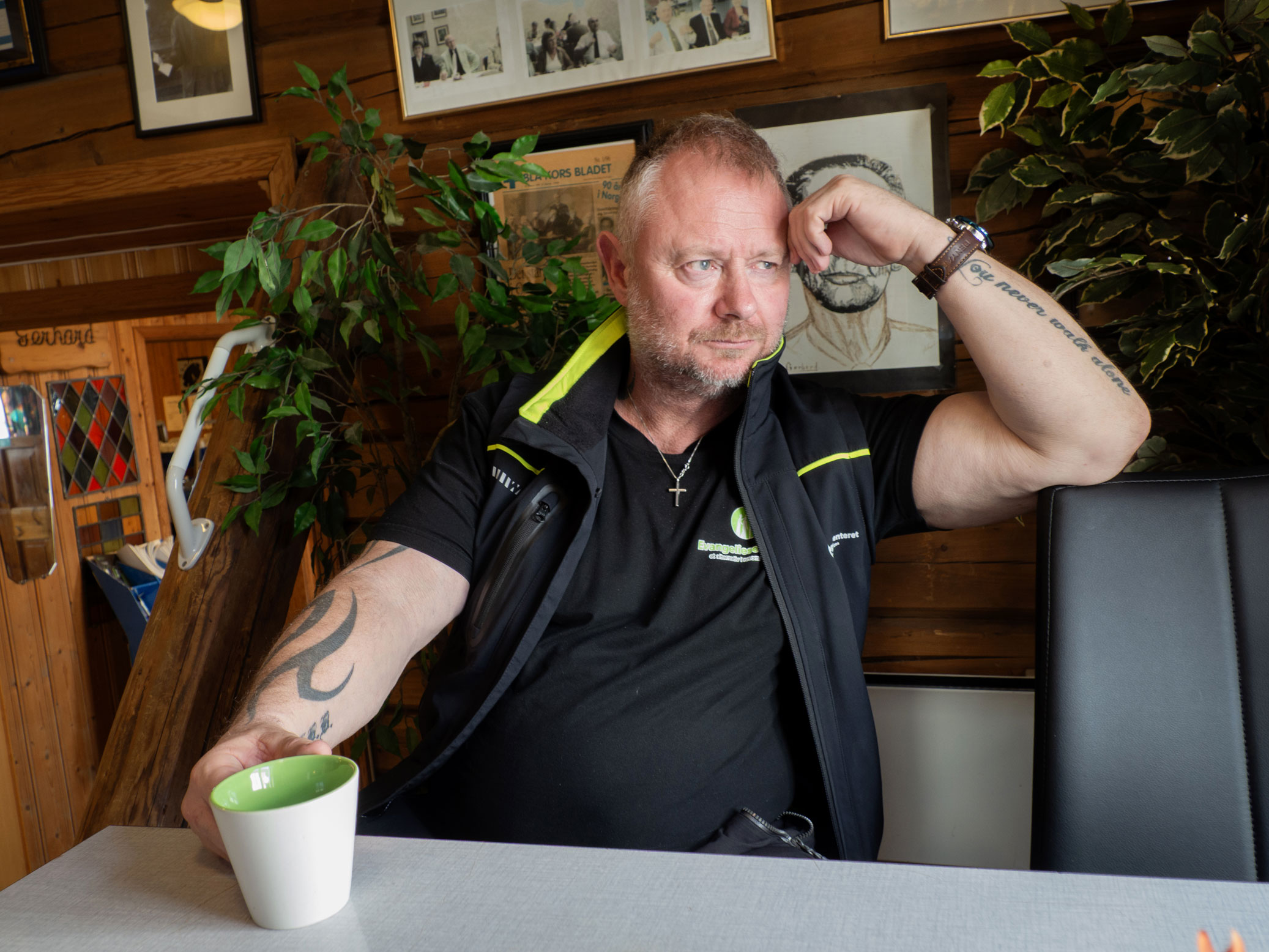Foto: Mann i 50-årene med kort hår og tatoveringer sitter ved et bort på en kafe. Han ser til siden.