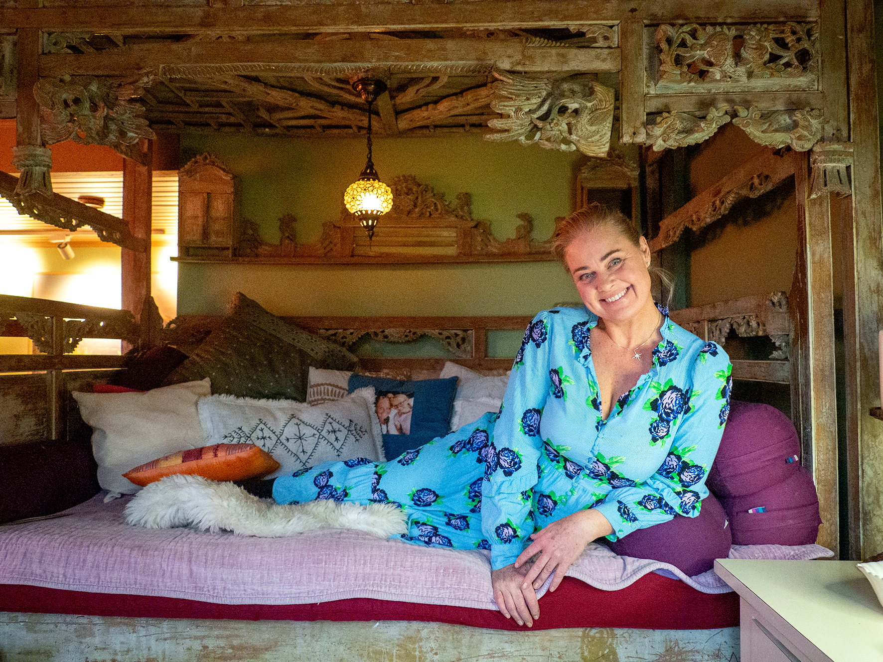 Foto: Kvinne ligger på en himmelseng fra østen med mange utskjæringer. Hun har på seg blåturkis kjole med blomster på.