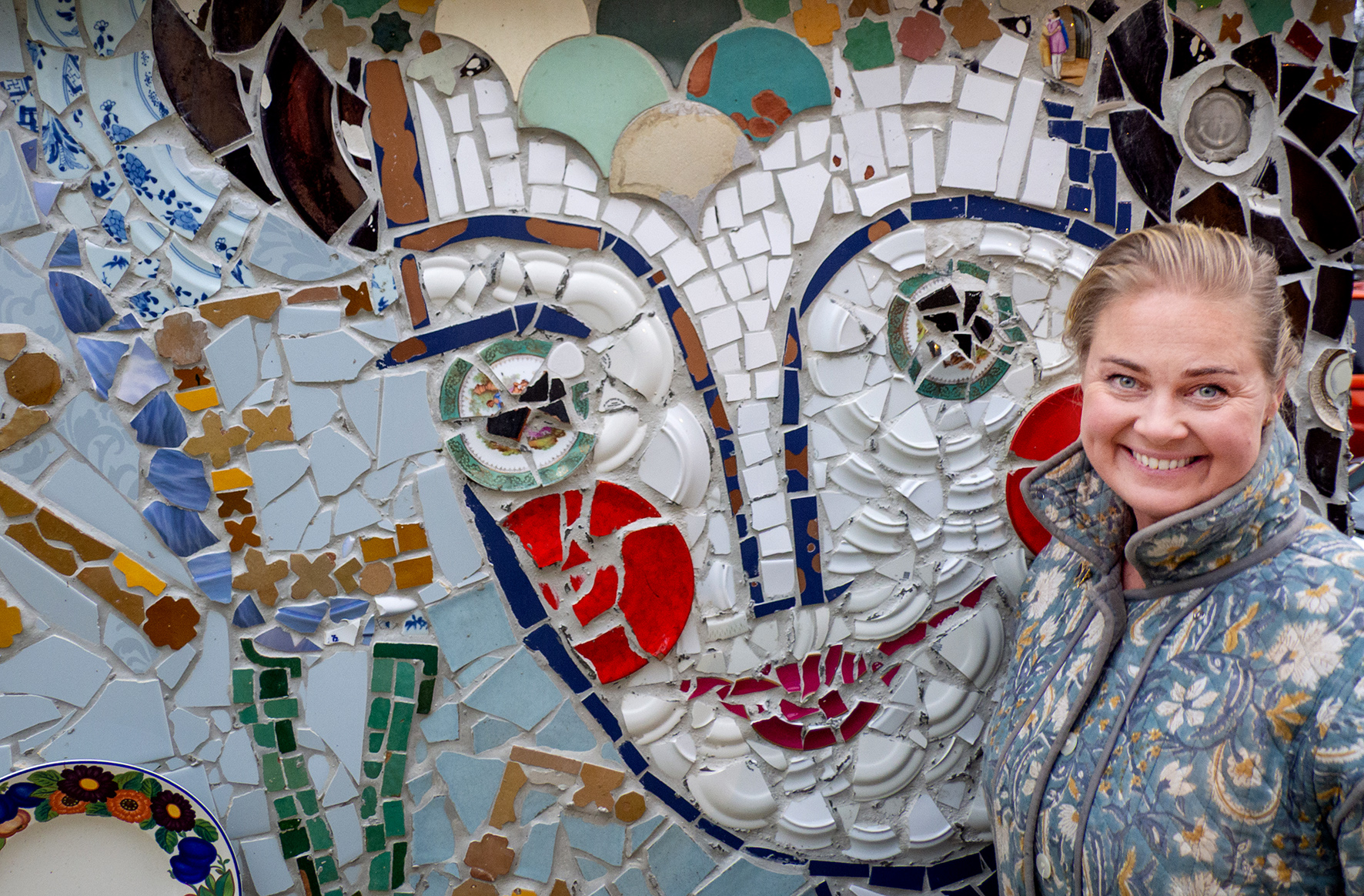 Foto: Blond kvinne med gråblå blomstrete kåpe står foran en vegg laget av knuste keramikkbiter. Bitene forestiller et ansikt.