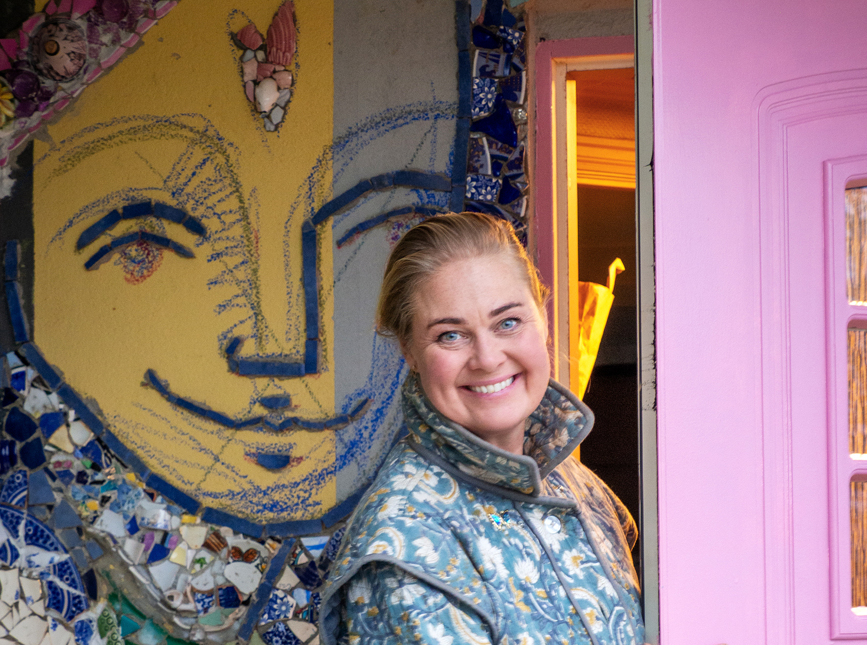 Foto: Smilende kvinne i 40-årene står midt i en døråpning. Hun har rosa ytterdør og laget mosaikk av et stort, smilende ansikt på veggen ved siden av. Hun har det lange, blonde håret oppsatt og har på en blomstrete kåpe i blågrå toner.