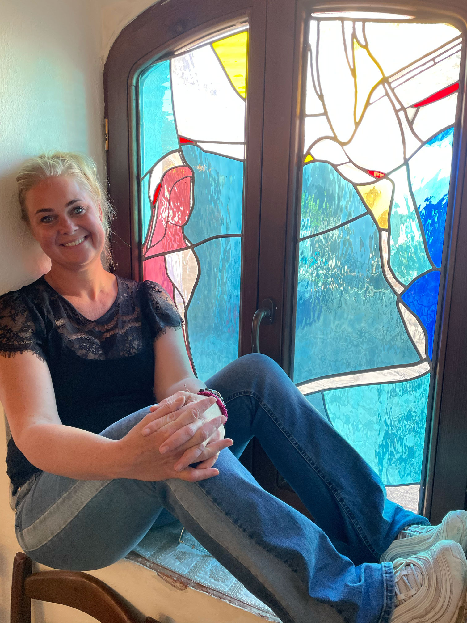 Foto: Foran et glassmaleri i et vindu sitter en blond, smilene kvinne i 40-årene med svart topp og jeans og hvite joggesko. Håret er oppsatt og vinduet viser en hvit fugl som kommer ned fra himmelen og mot en kvinneskikkelse.