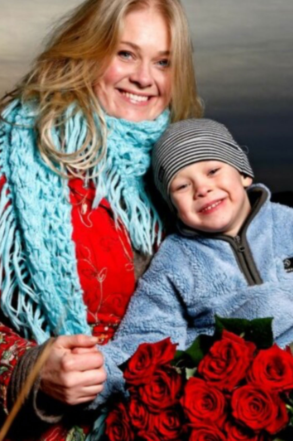 Foto: Ung kvinne med langt, blondt hår i en rød kåpe og turkis skjerf. Hun er sammen med en liten gutt i blå flisjakke og lue. Begge smiler og de holder en bukett med roser.