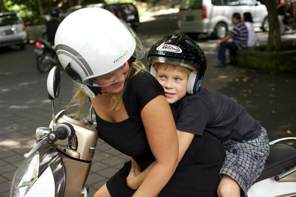 Foto: Kvinne med hvit hjelm kjører en moped med en ung gutt med svart hjelm.