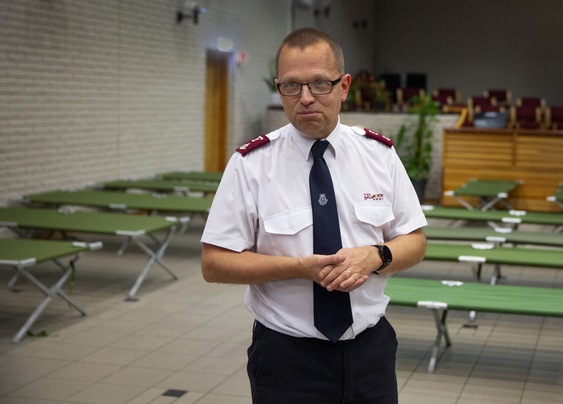 Foto: Mann i hvit skjorte fra Frelsesarmeen står i et kirkelokale med en rekke grønne feltsenger oppslått over alt i det store rommet. Han er i 50-årene og har kort, mørkeblondt hår og briller.