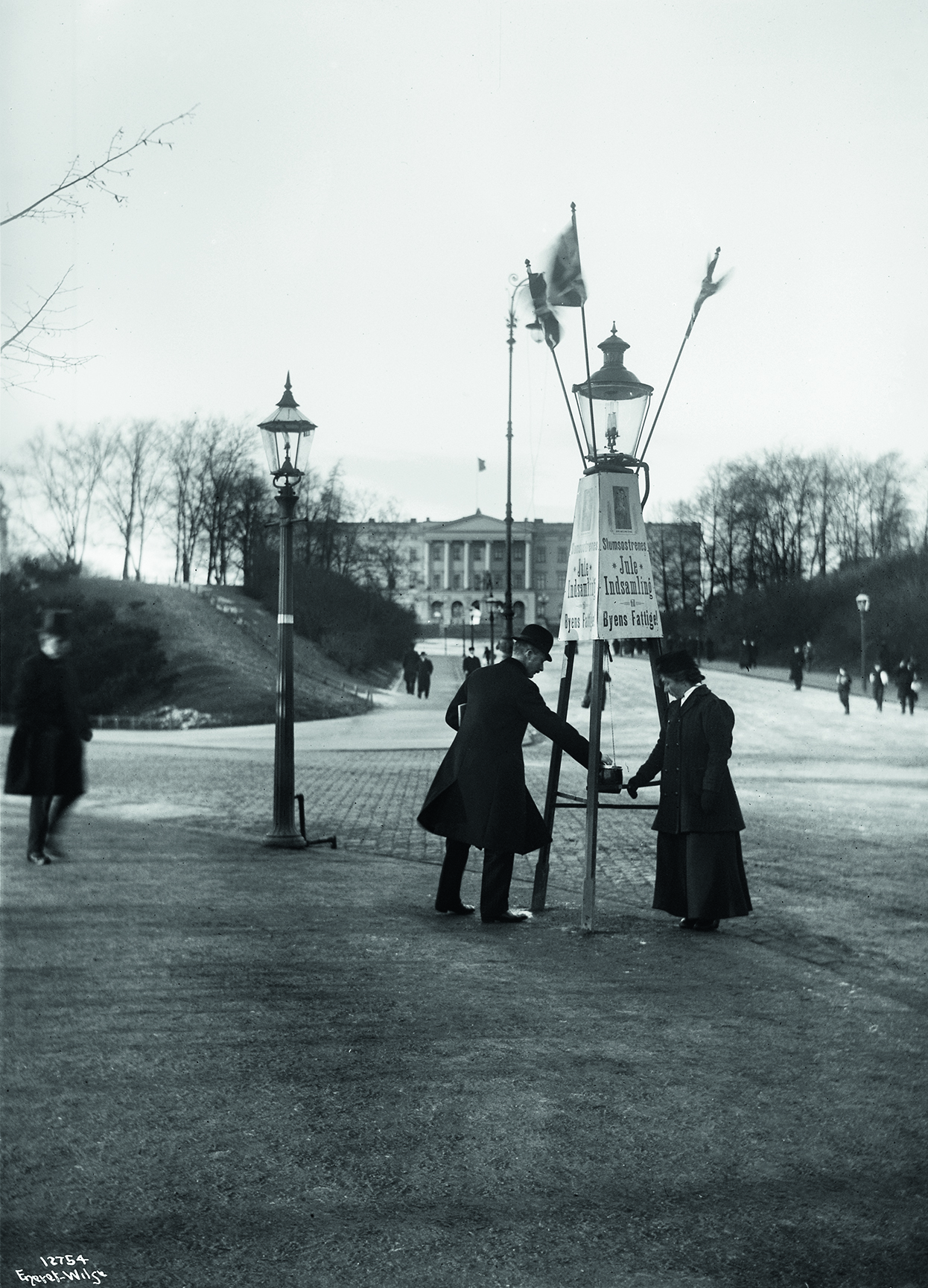 Gammelt bilde i sort hvitt av Frelsesarmeens julegryte med folk i klær fra starten av 1900-tallet. Stativet med gryta står øverst på Karl Johan med slottet tronende i bakgrunnen.