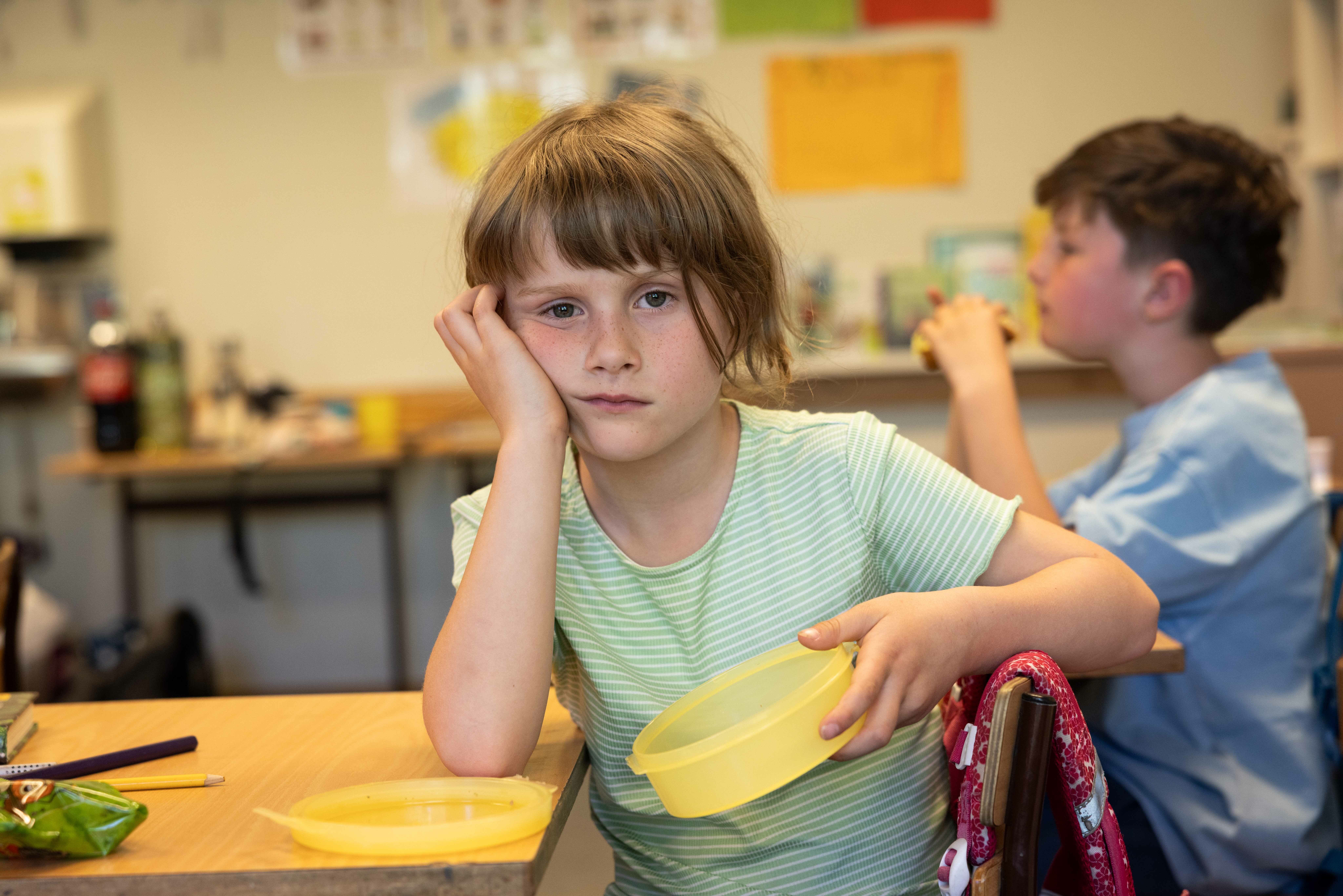 En jente i et klasserom med tom matboks ser oppgitt ut i kameraet. Rundt henne sitter klassekamerater med mat i matboksen. De spiser.