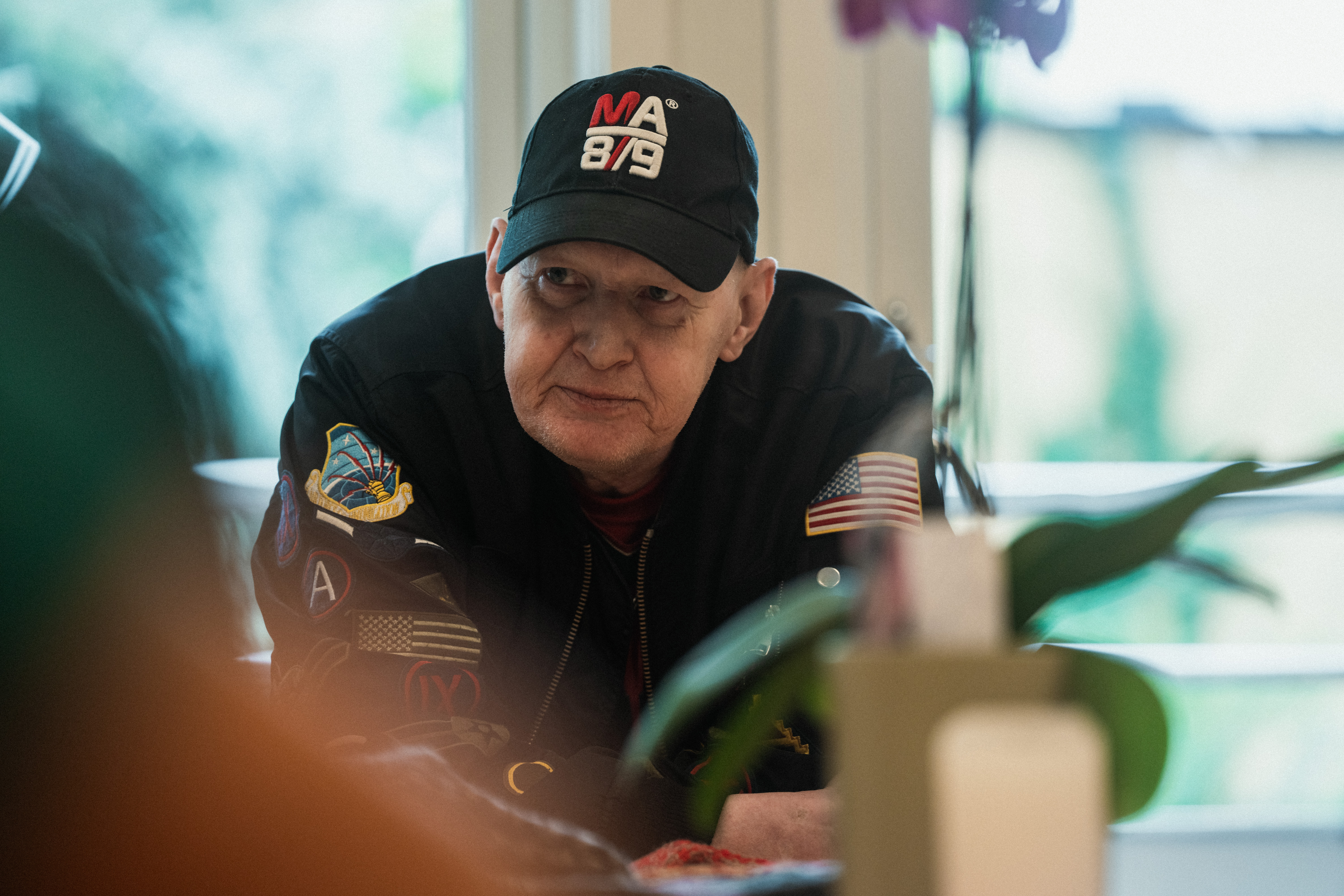 Vi ser Bjørn som sitter ved et bord. Han har svart jakke og svart caps med merker og bokstaver. Han snakker og kikker med blikket ut i luften.