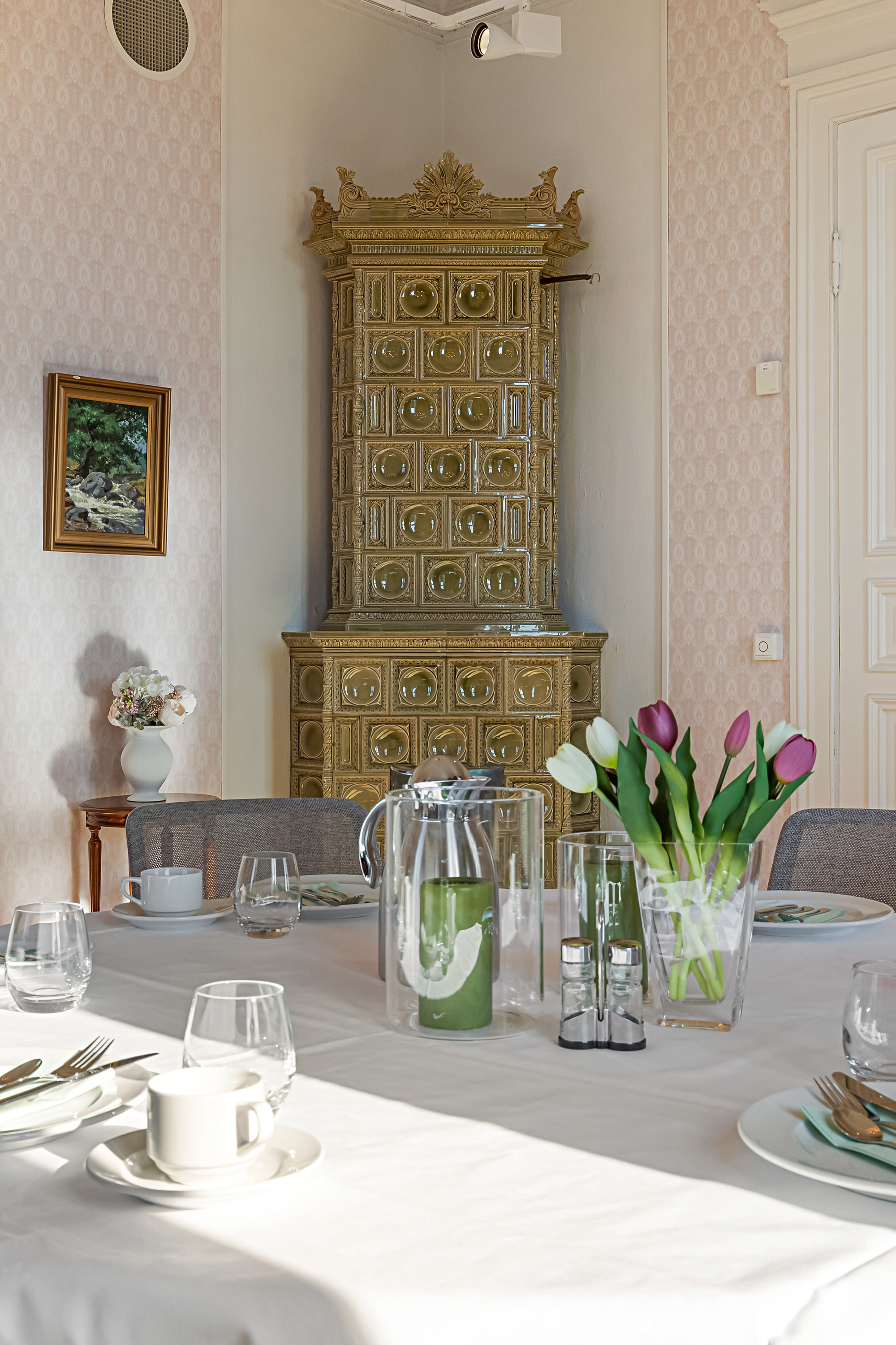 Bilde 1: Nærbilde av et oppdekket bord med tulipaner og lys på bordet, i bakgrunnen er det en høy gammel grønn kakkelovn. Bilde 2: Rom med gammeldags rosa tapet, et pent oppdekket rundt bord med kaffekopper, kaffekanne, tulipaner og lys. I bakgrunnen er det en gammel trefarget bokhylle og et høyt trefarget speil langs veggen.