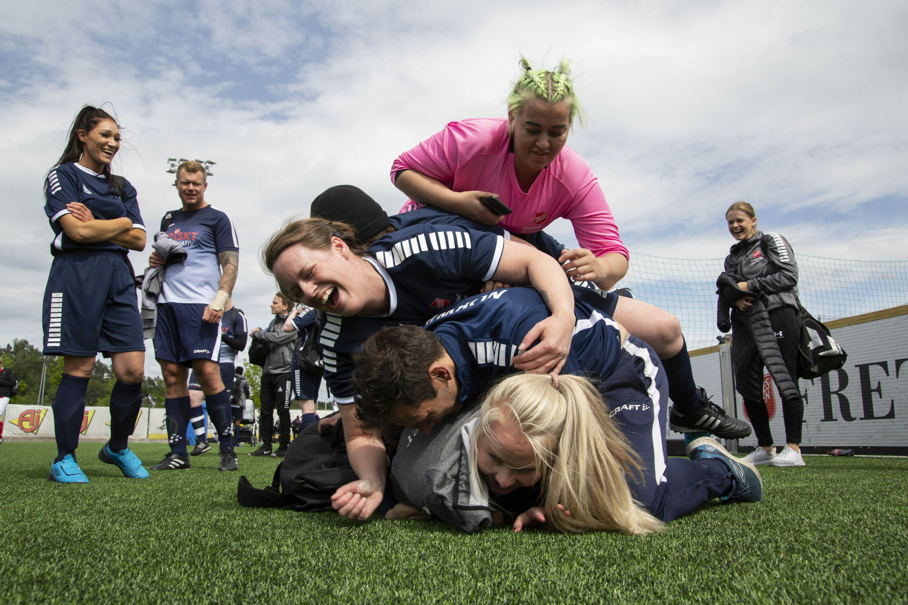 Foto: Fire fotballspillere, tre kvinner og en mann, ligger slengt over hverandre i en "haug" på gressmatta. De ler godt. Rundt dem står flere andre spillere og smiler.