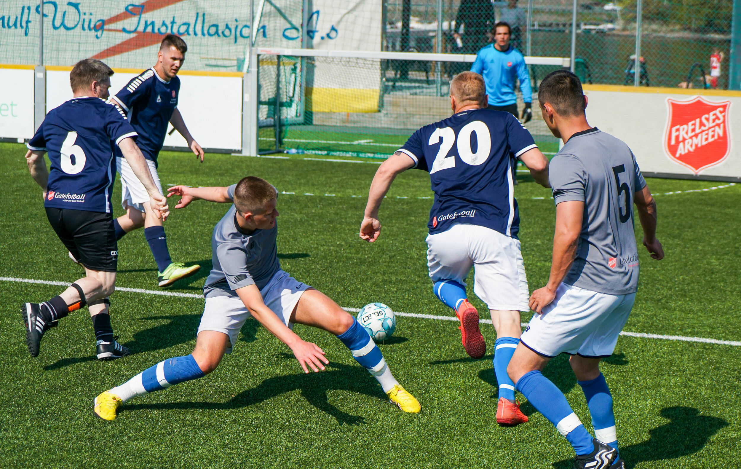 Foto: Fem menn i fotballdrakter som kjemper om ballen på en bane. I mål følger keeper godt med.
