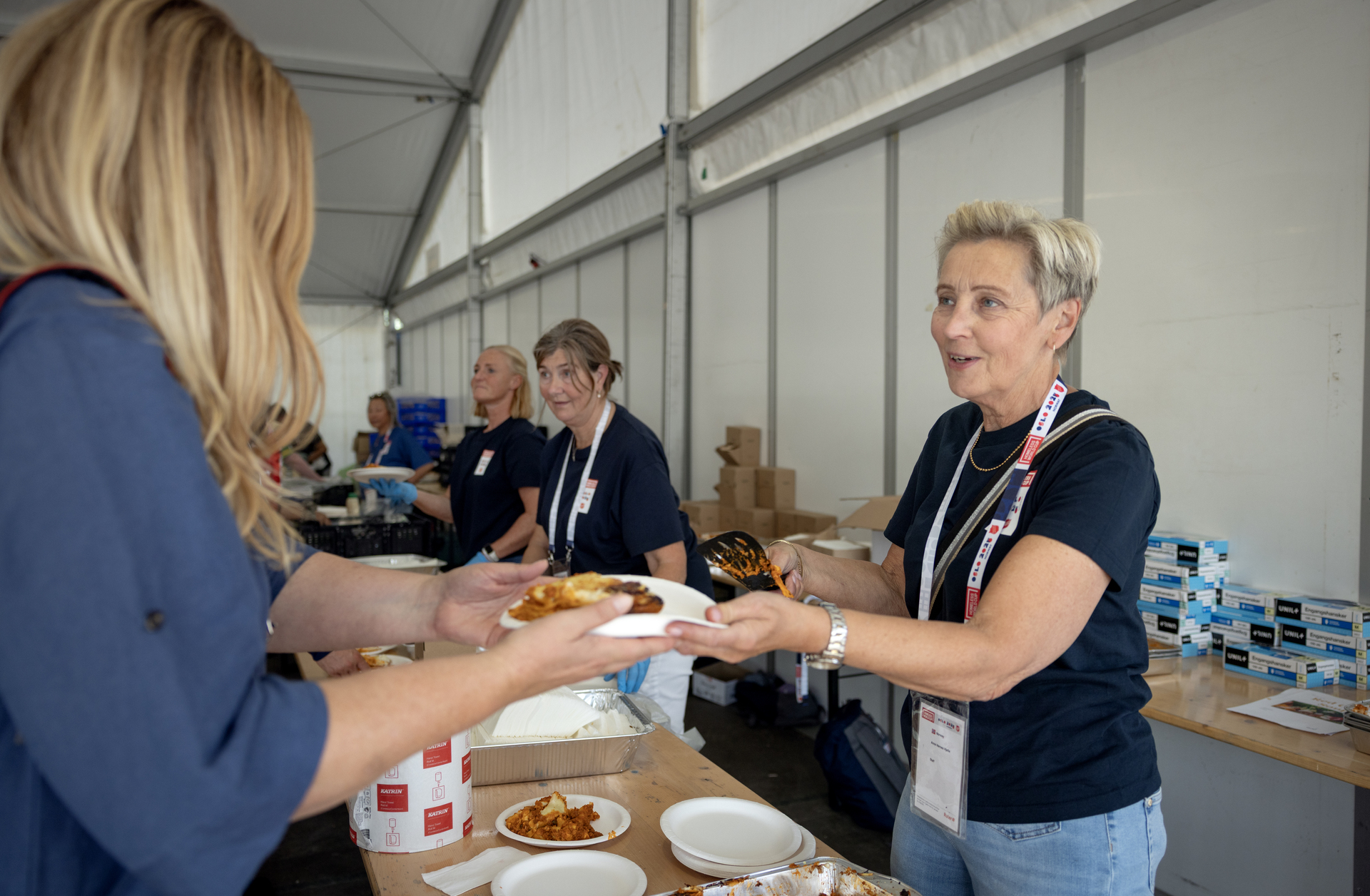 Tre damer står bak et langbord og serverer middag til deltakere på Homeless World Cup.