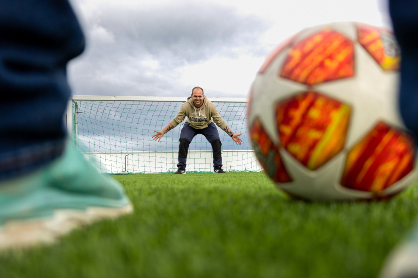 Foto: Mann står klar i mål. Krøket sammen. Nær kamera ligger en fotball (den er da stor) og man ser litt av en fot med grønn joggesko. Frelsesarmeen gatefotball.