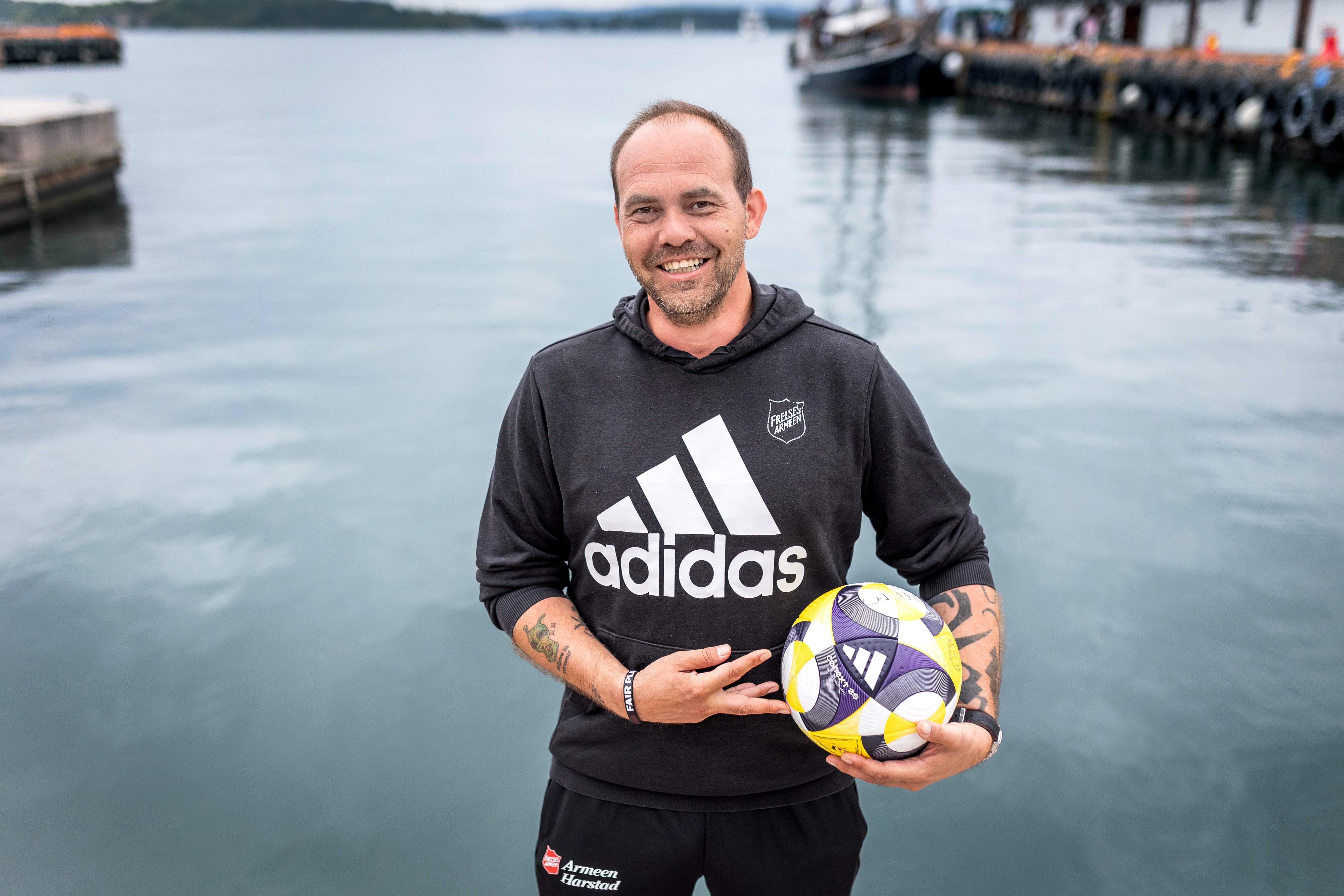 Foto: En mann i svart Adidas-genser med Frelsesarme-logo står ved vannet på Rådhuskaia og smiler. Han holder en fotball i den ene armen.