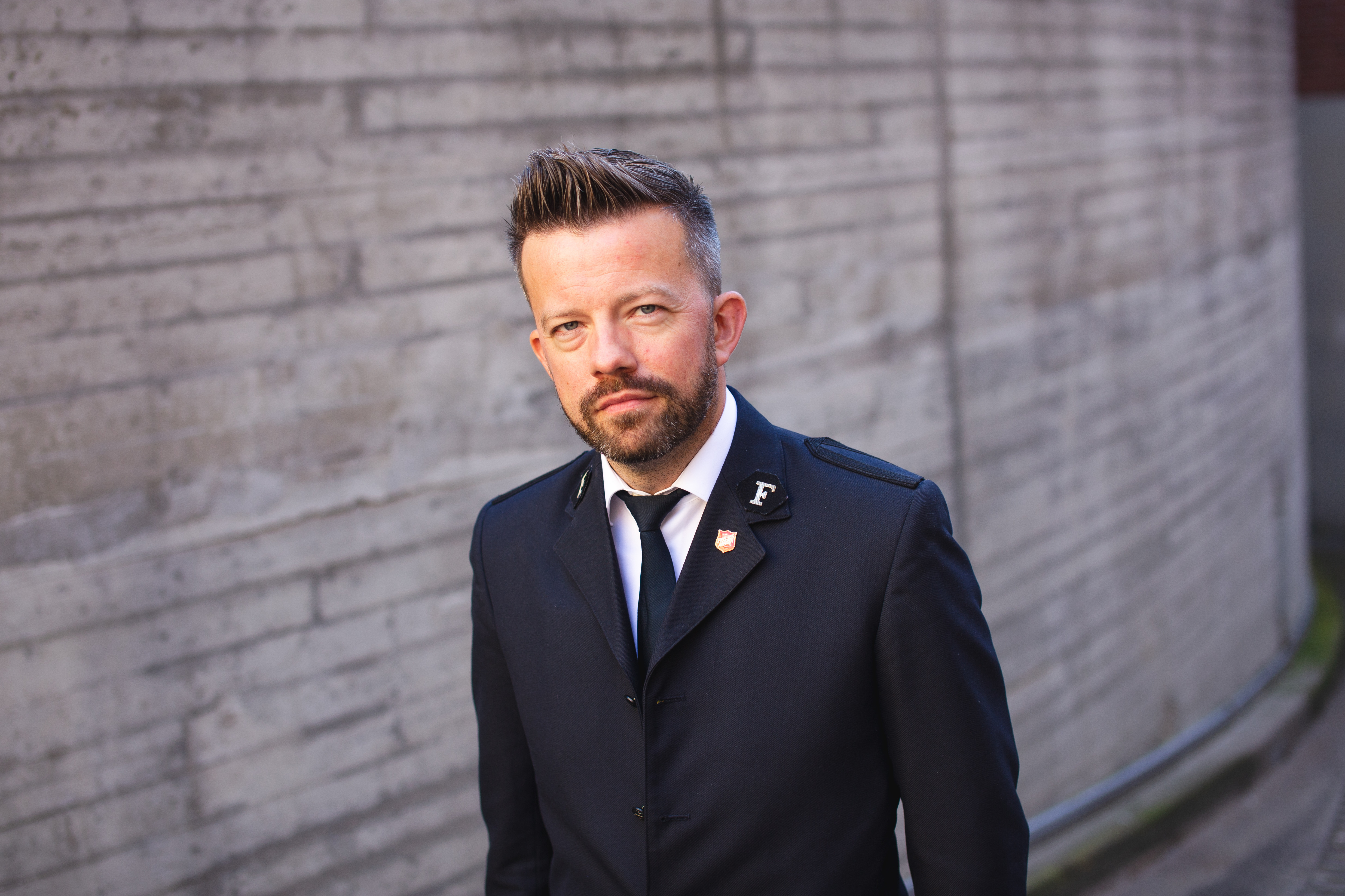 Foto: Mann i 40-årene med kort hår og skjegg i uniform fra Frelsesarmeen. Han står foran en murvegg ute.