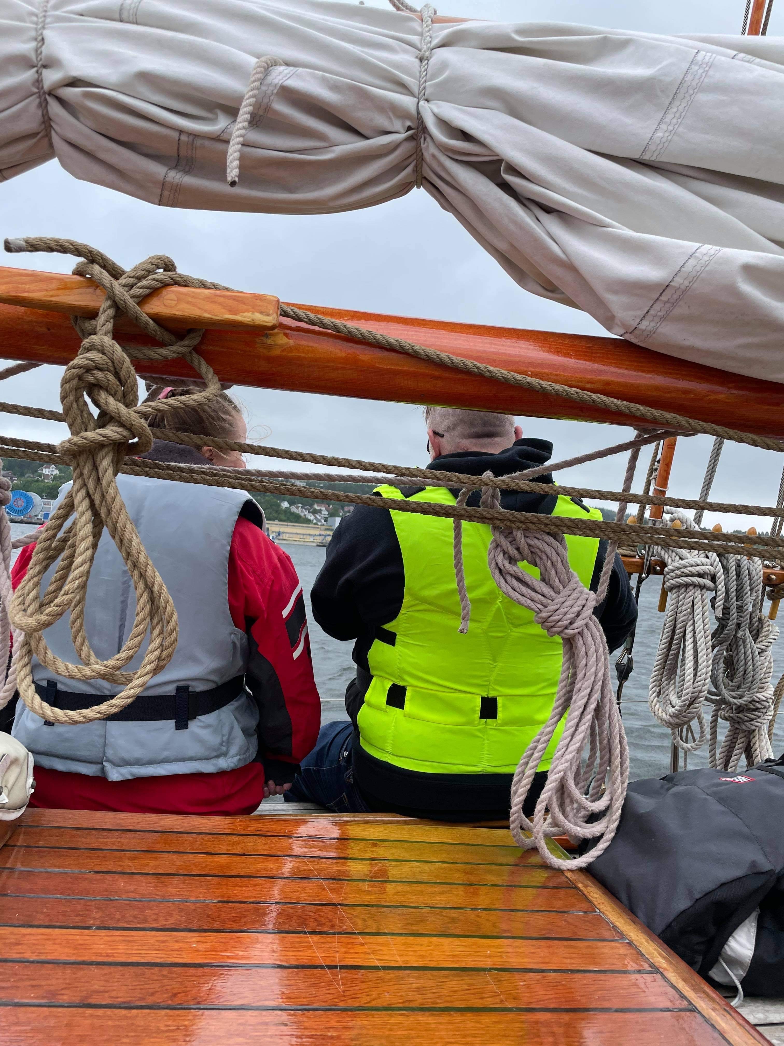 To som sitter med ryggen til på en seilbåt. De har redningsvester på.