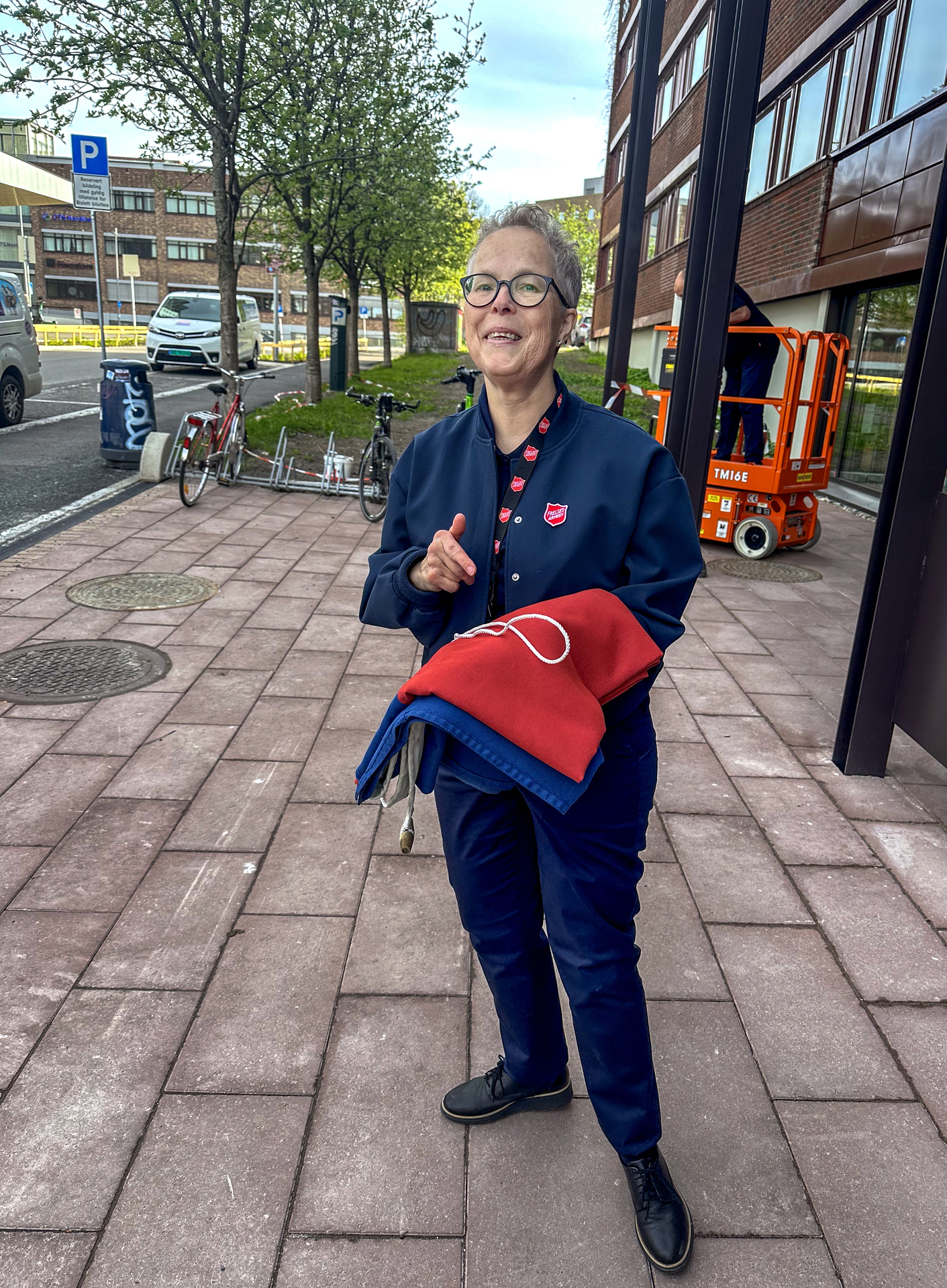 Foto: Kvinne i 60-årene med kort hår og briller står utenfor en bygning med et sammenbrettet flagg. I bakgrunnen sees en oransje lift.