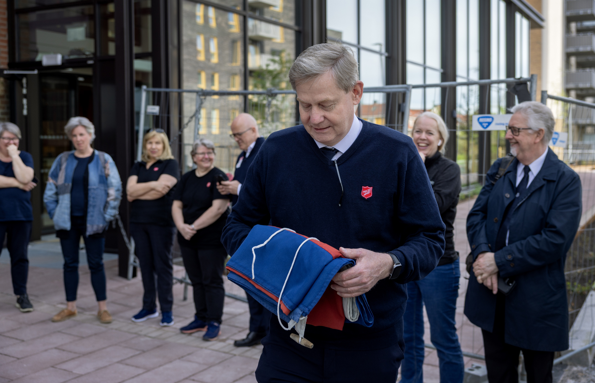 Foto: Mann i 60-årene har et sammenbrettet flagg liggende over høyre arm. Han ser ned på det. Bak ham står en liten gjeng med folk, stort sett i profiltøy fra Frelsesarmeen.