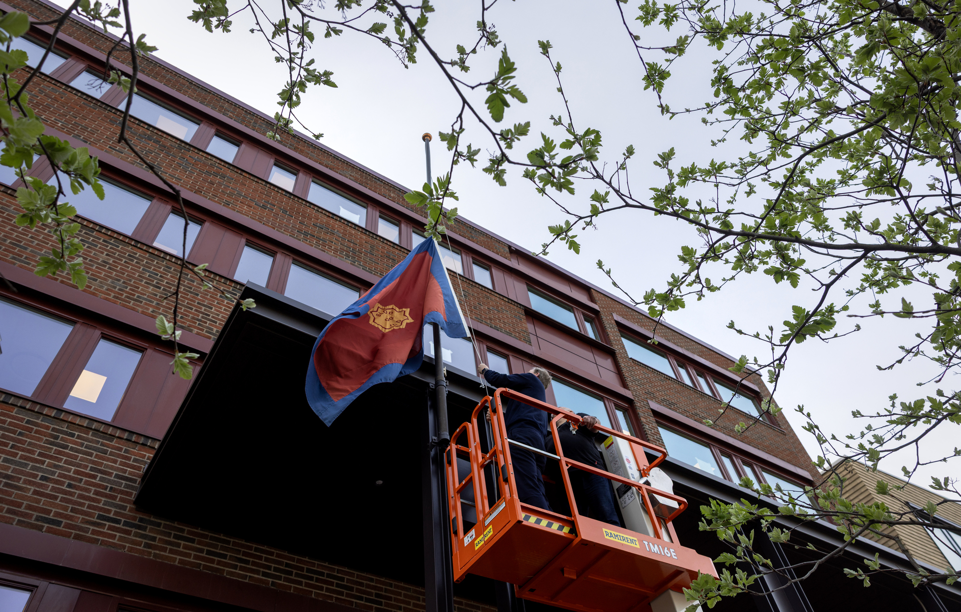 Foto: Flagg i ferd med å heises opp fra en person som står trygt i en boks oppe i en oransje lift.