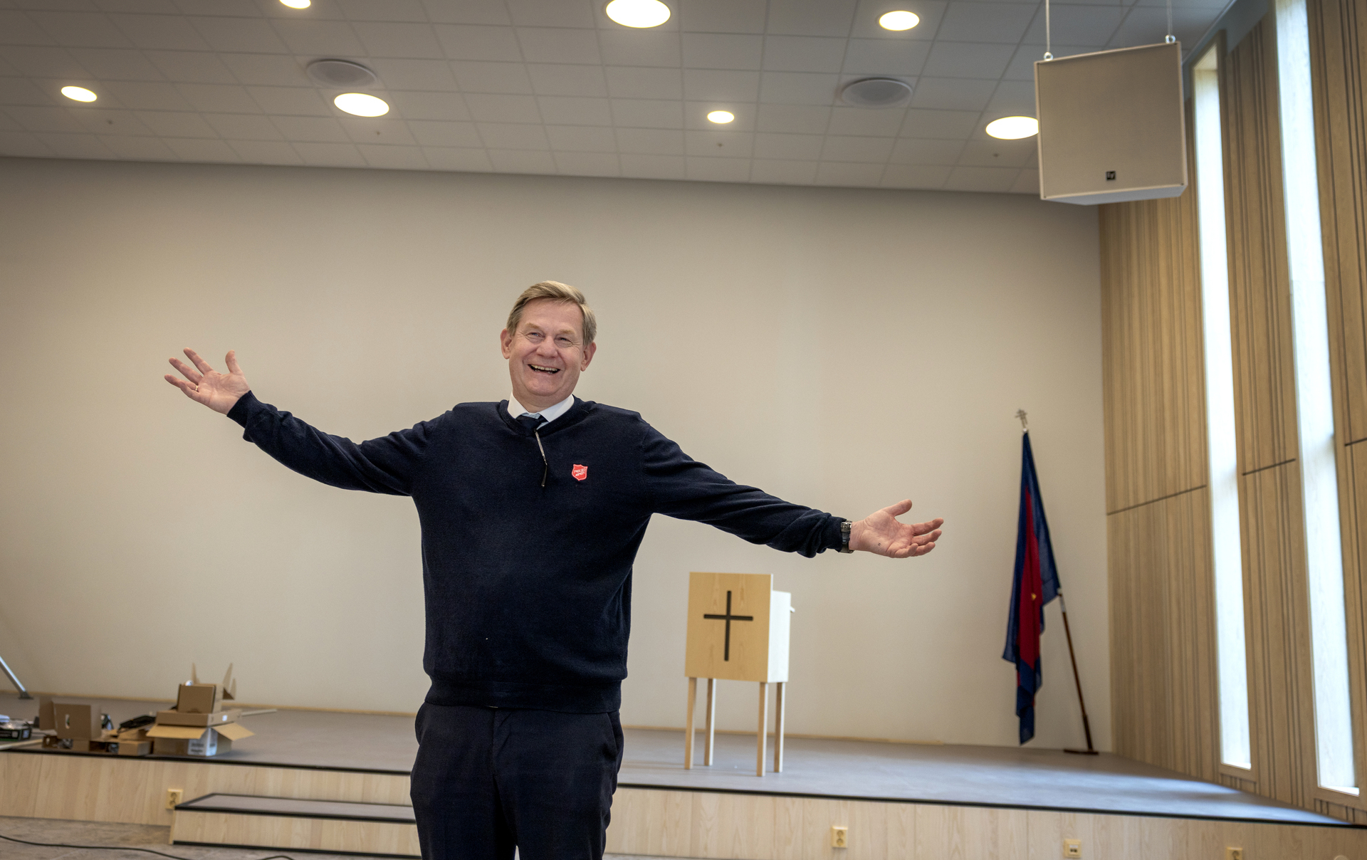 Foto: Mann med utstrakte armer i et stort kirkerom med en enkel talerstol med kors på og et likt flagg som det utenfor stående i hjørnet. Det er tydelig at ting blir montert i rommet