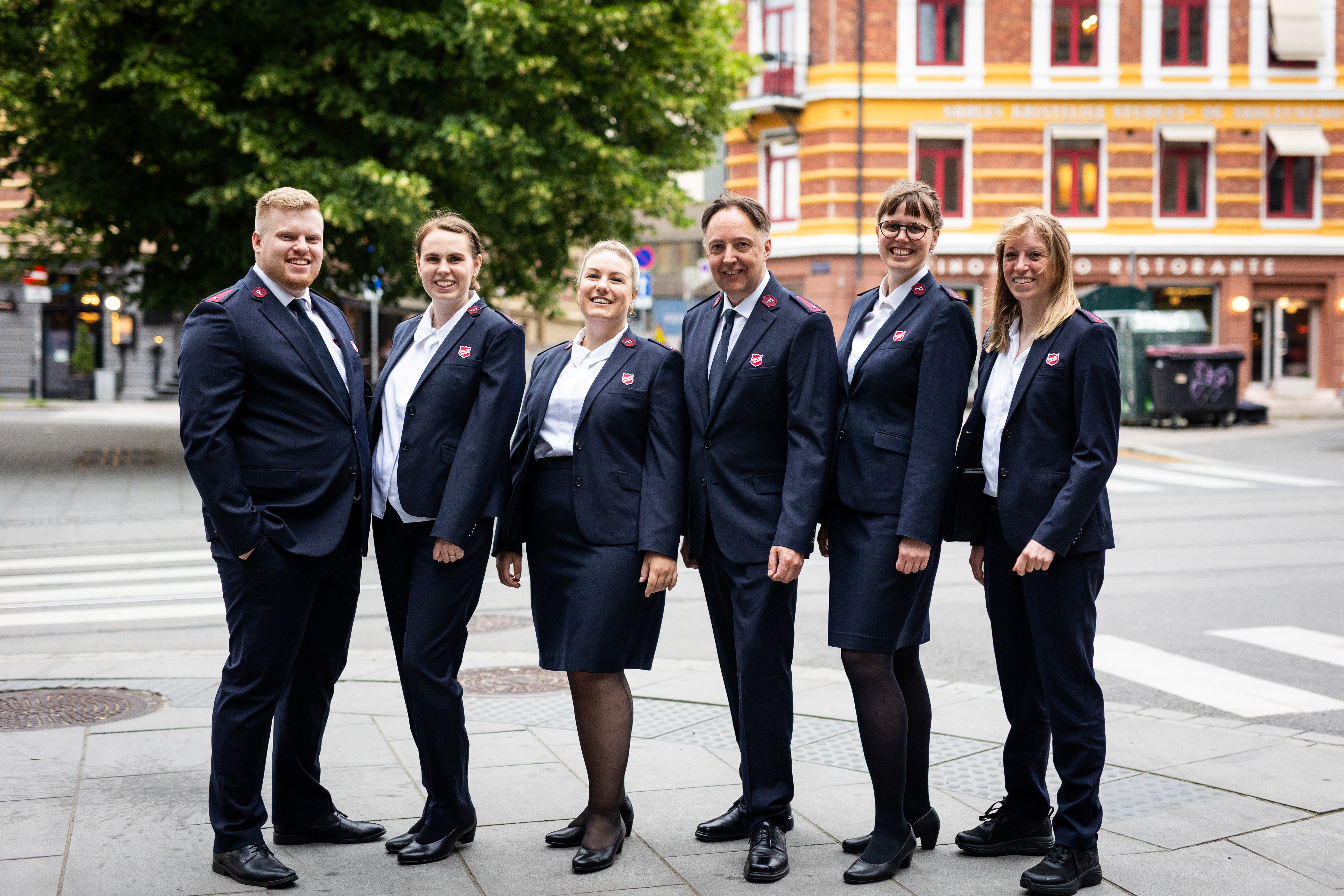 Foto: To menn og seks kvinner i uniformer fra Frelsesarmeen står på en rekke i bybildet.