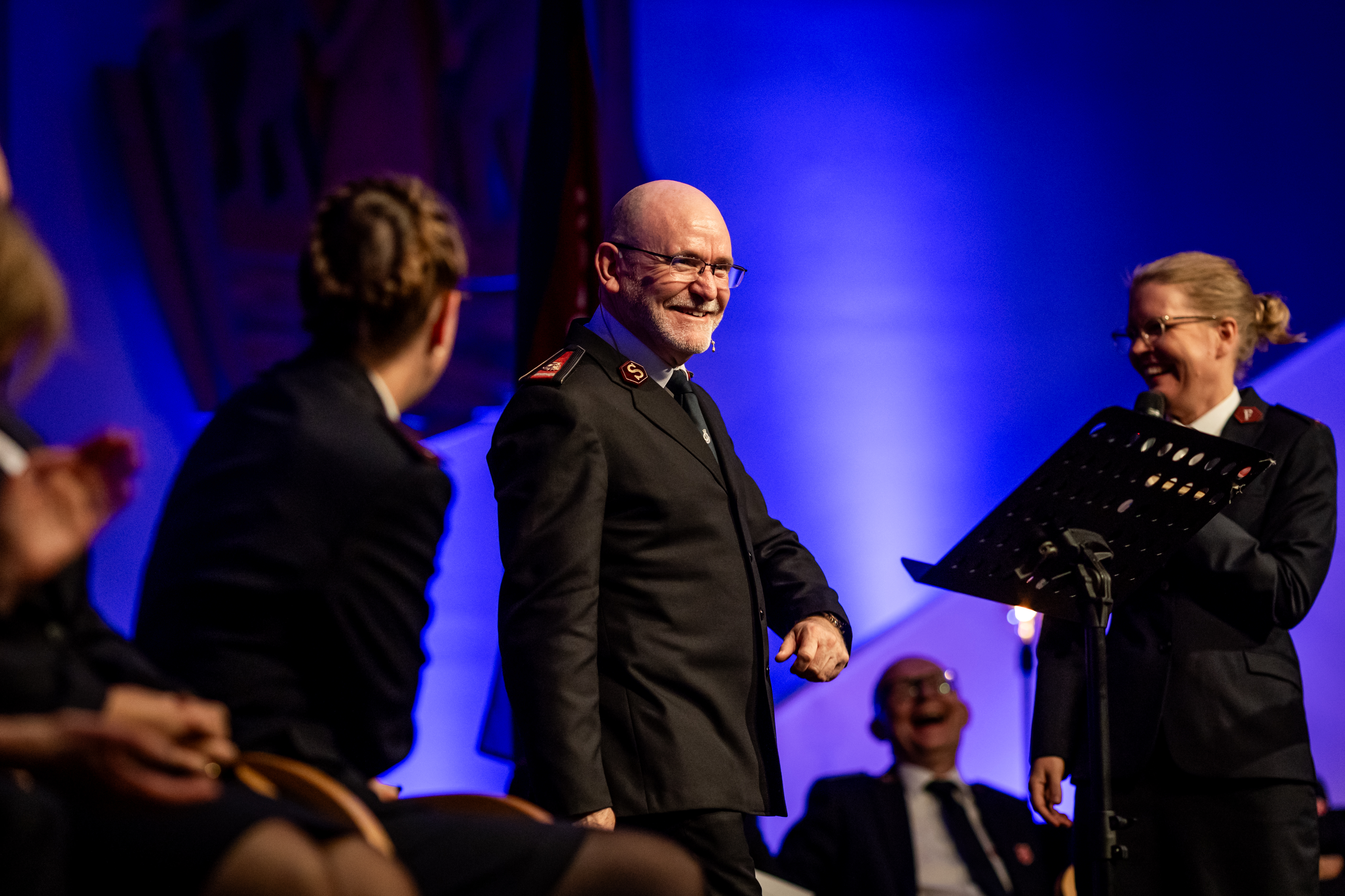 Vi ser General Lyndon Buckingham på en scene. Flere mennesker på samme scene ler ham fordi han traff både hjertet og lattermusklene under sin tale. Tolk Janne Våje Breen til høyre.