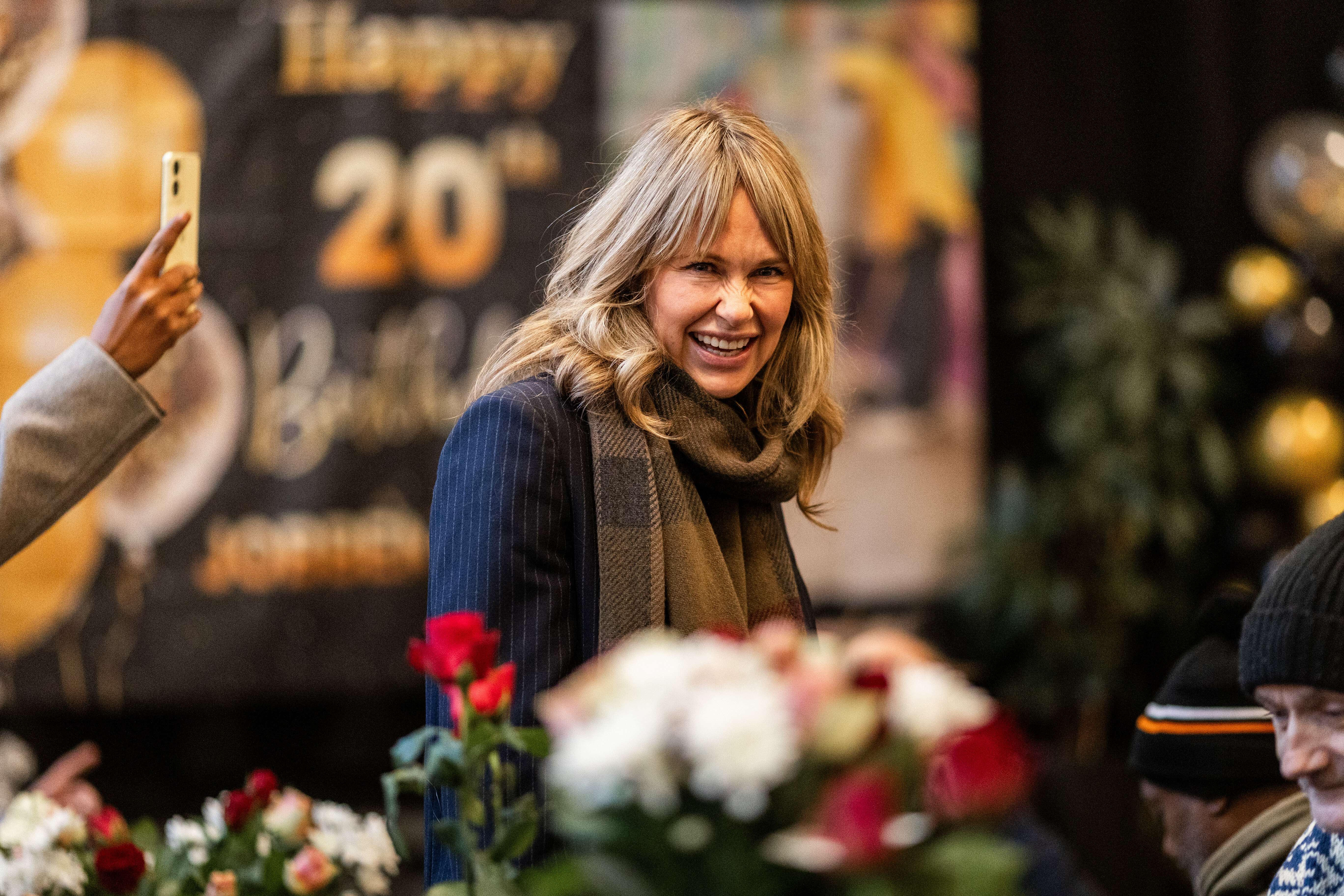 Foto: Blond kvinne i 50-årene med hår ned til skuldrene smiler til fotografen. På veggen bak henne henger en plakat der det står tallet 20. Foran er et bort med planter og julepynt.