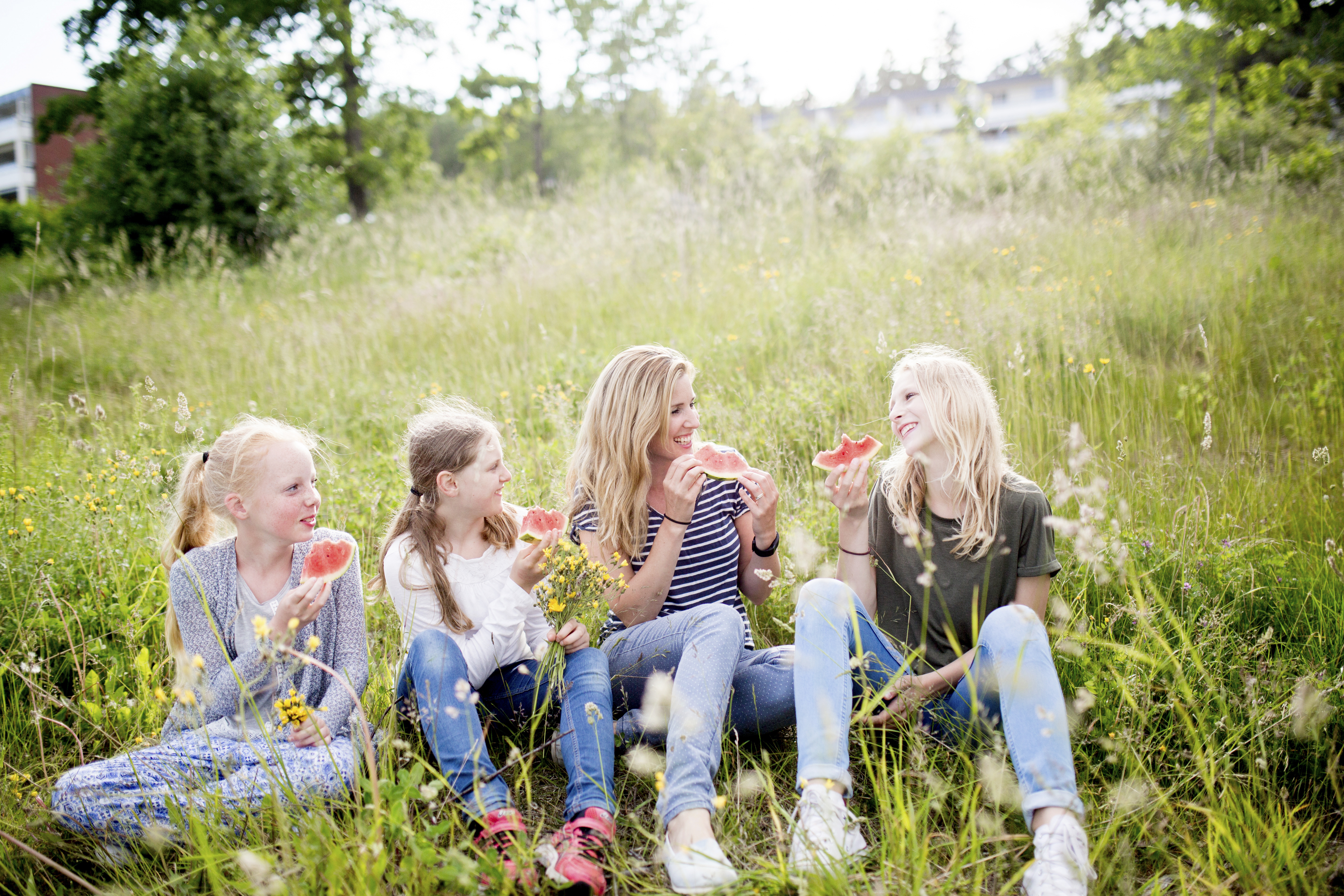 4 jenter i ulik aldre sitter i blomstereng og smiler og spiser vannmelon