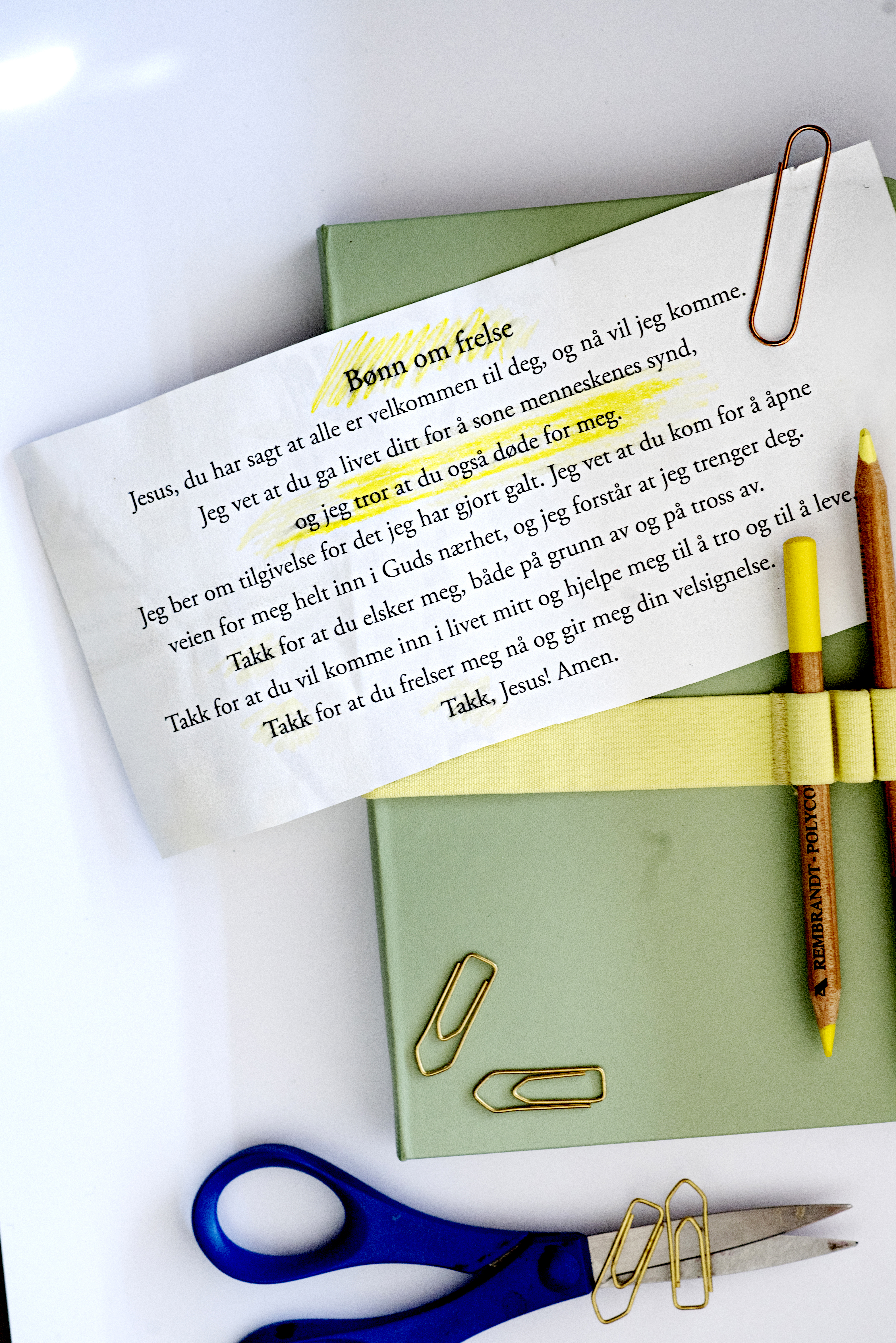 Bilde av en papirlapp med en bønn om frelse. Lappen ligger oppå en grønn notatbok og binderser ligger litt lett strødd rundt. Papirlappen med bønnen har blitt skriblet litt på med en gul markeringspenn.