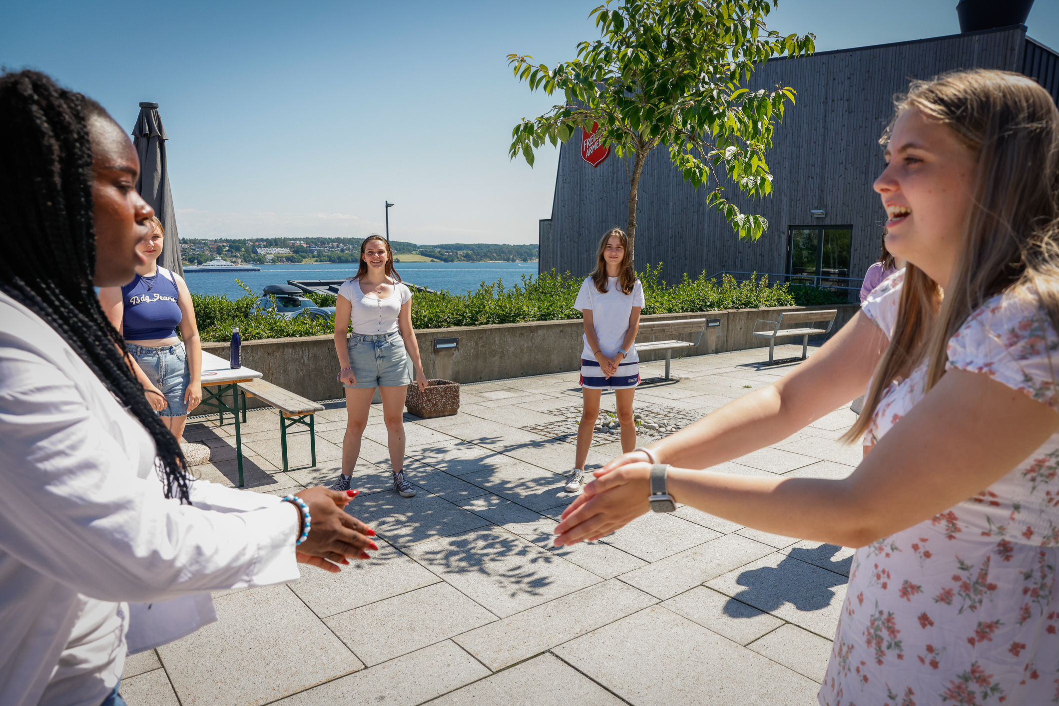 Seks jenter som står i ring ute i solskinnet. To av dem står mot hverandre og klapper i hendene.