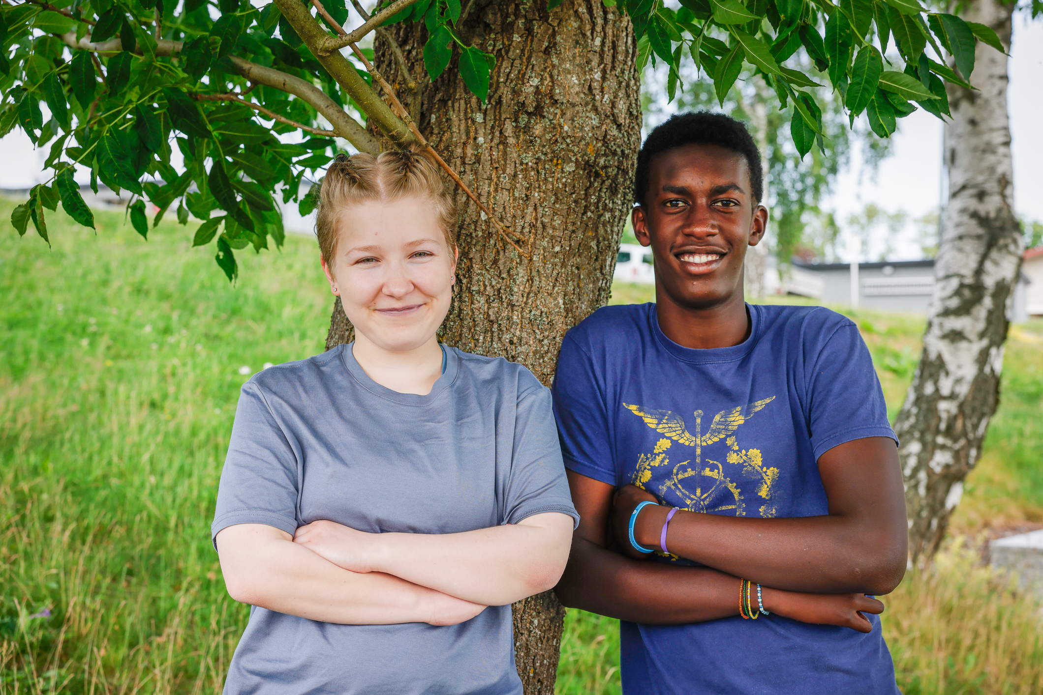 Tenårings jente og gutt står under et tre. Begge har armene i kors.