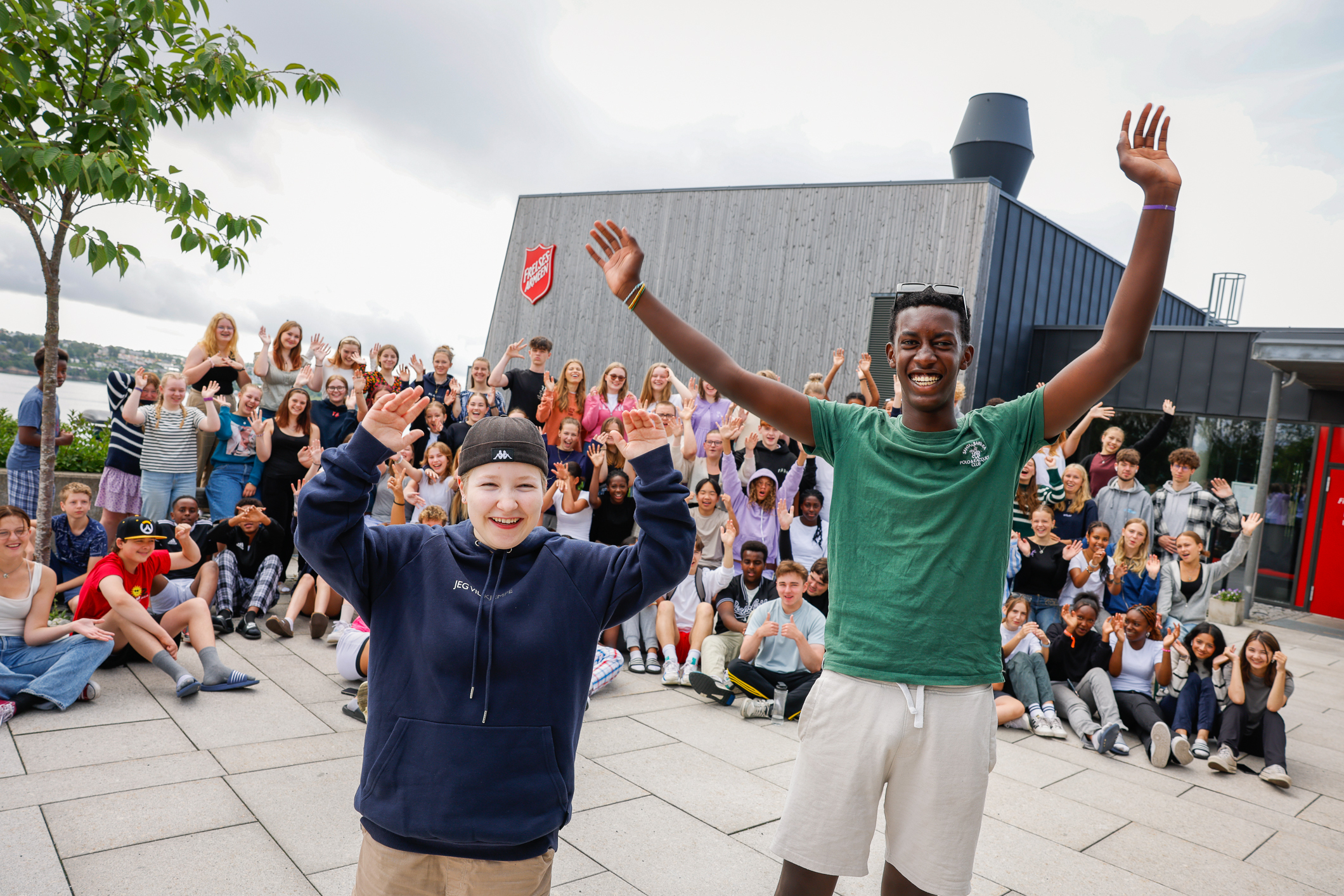 En hel haug med ungdommer på et slags lagbilde. De står og sitter foran et moderne bygg. Mange veiver med armene.