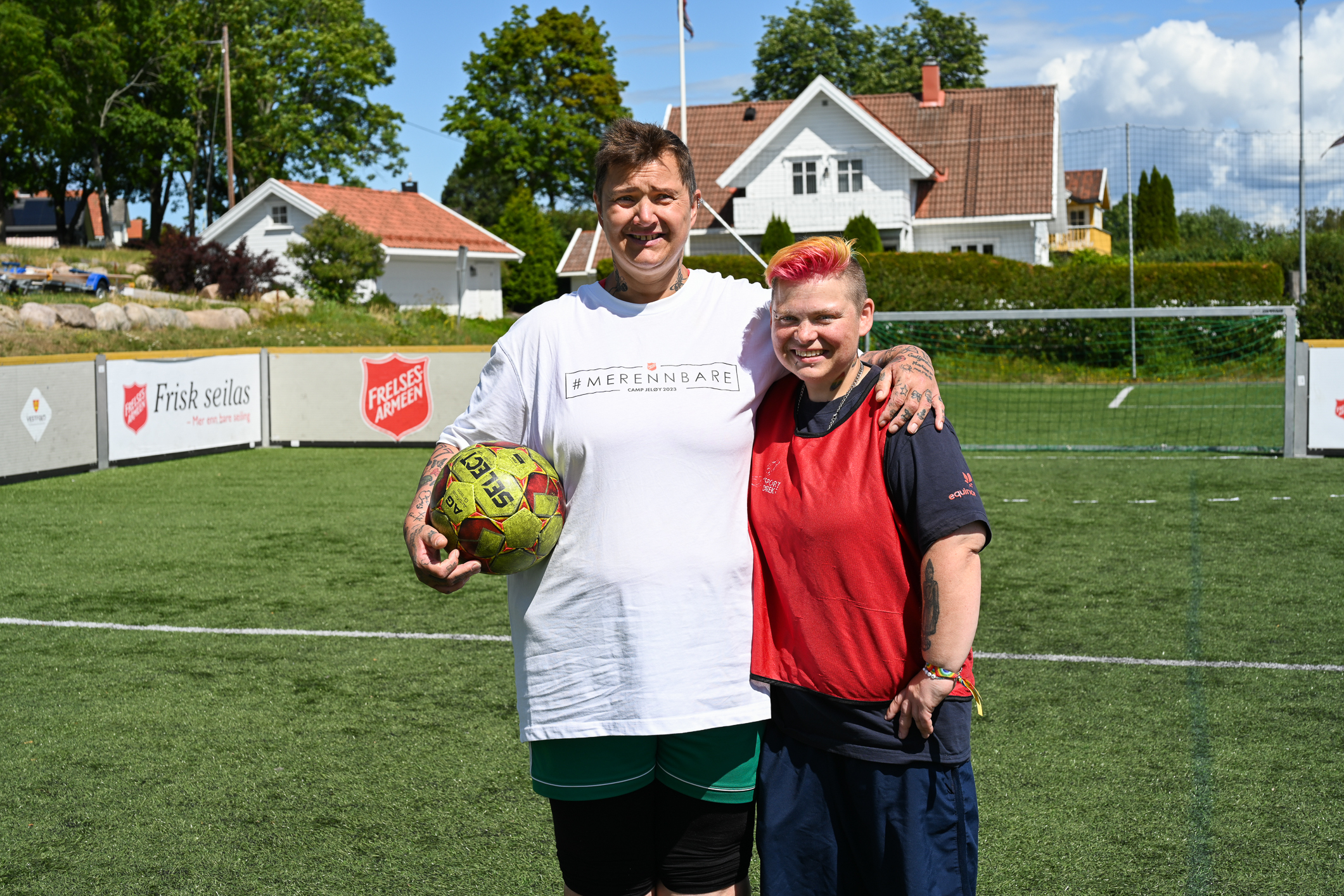 To kvinner står oppstilt på en grønn gressmatte med et mål i bakgrunnen. Den ene har en fotball under armen.