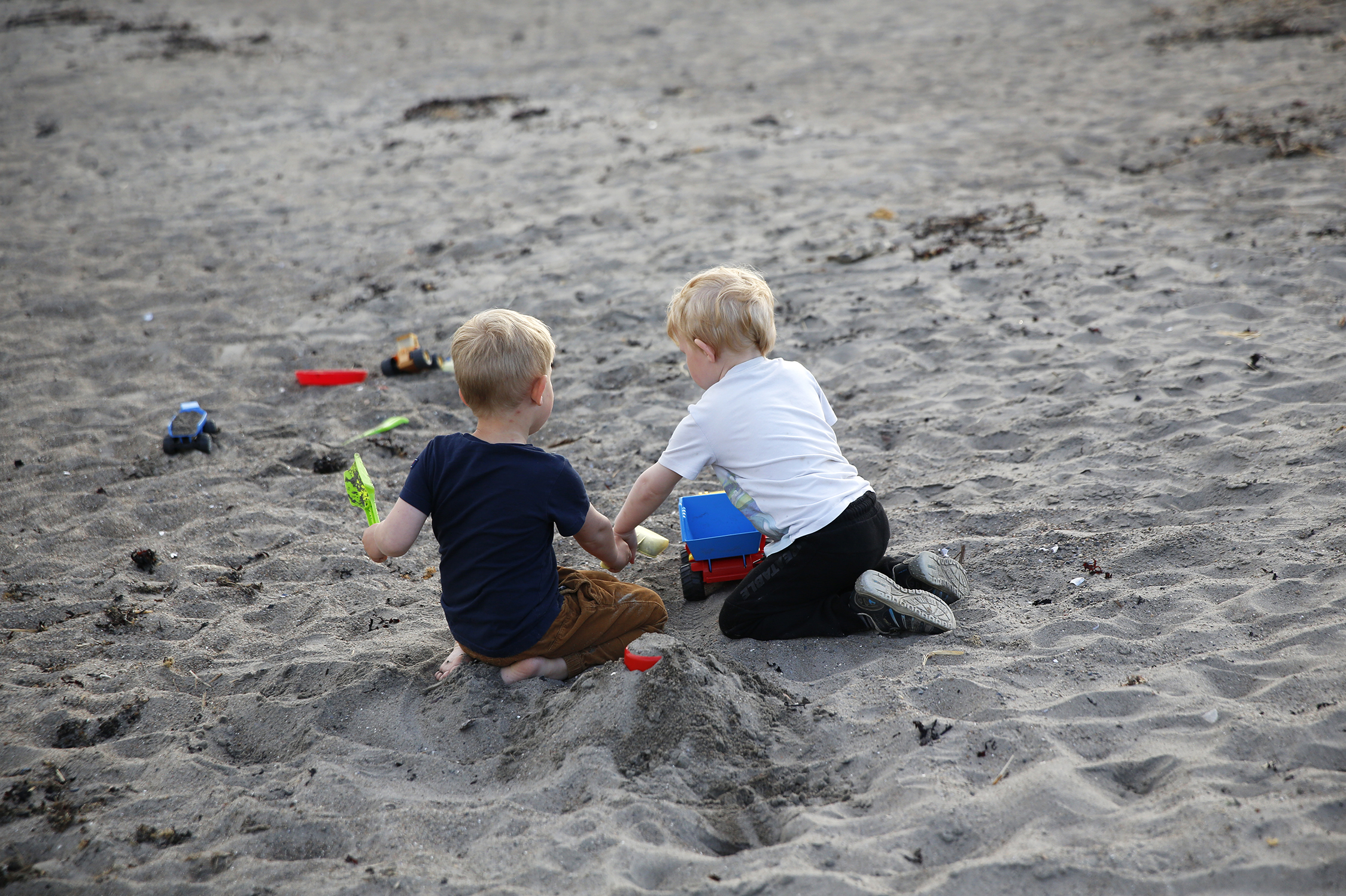Bildet viser to barn som leker på en sandstrand. De har ryggen til og  graver i sanden