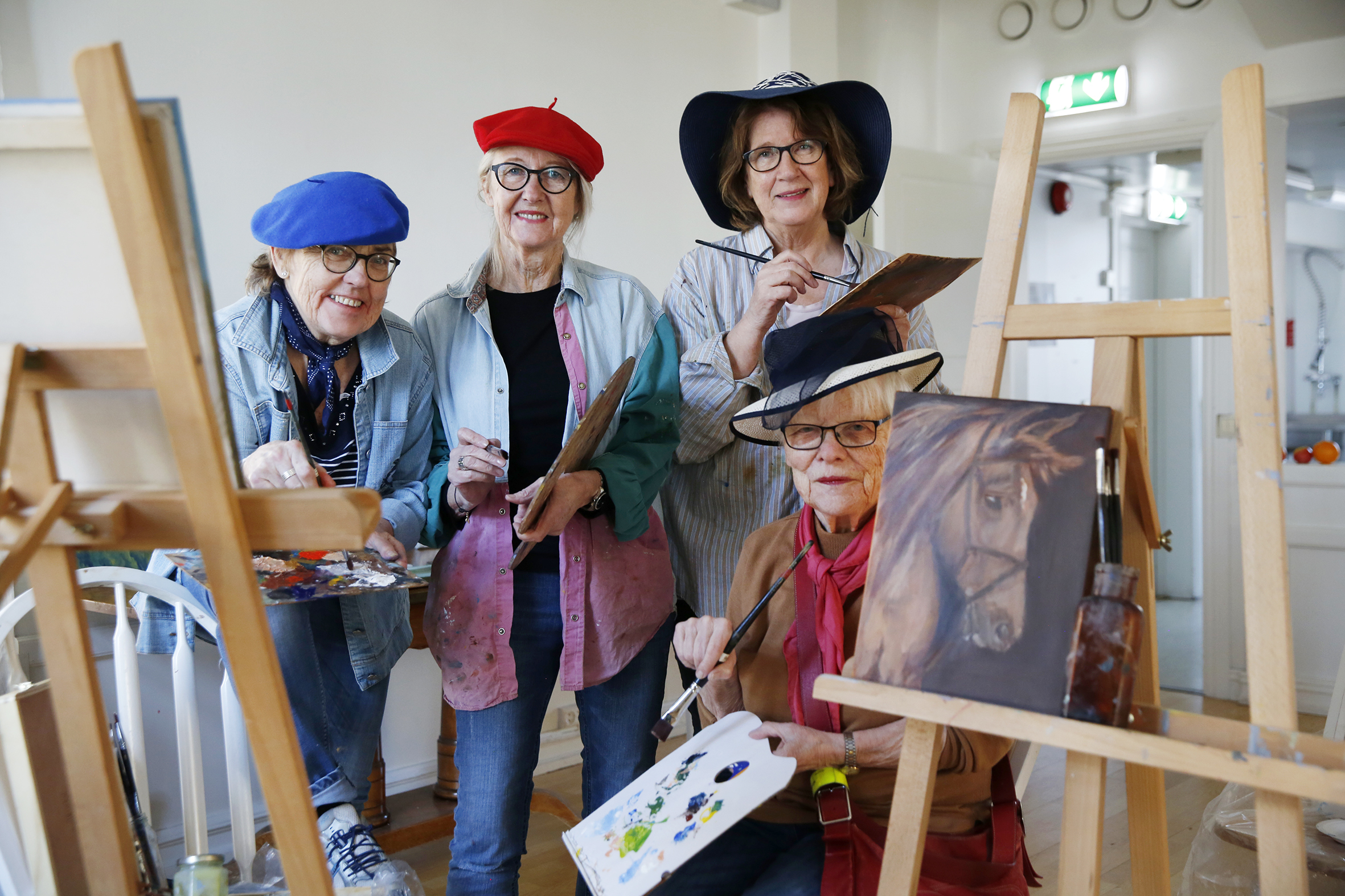 Bildet viser fire damer i fargerike kunstnerhatter. De sitter blant maleriene sine og holder hver sin palett.