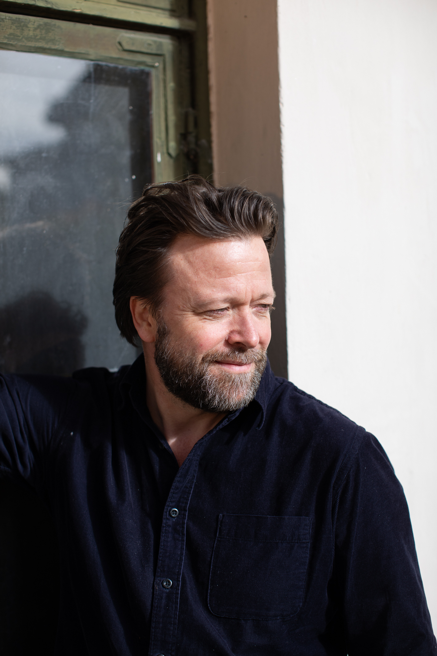 Mann som kikker litt til siden. Står ute foran et vindu med en grønnmalt karm.
