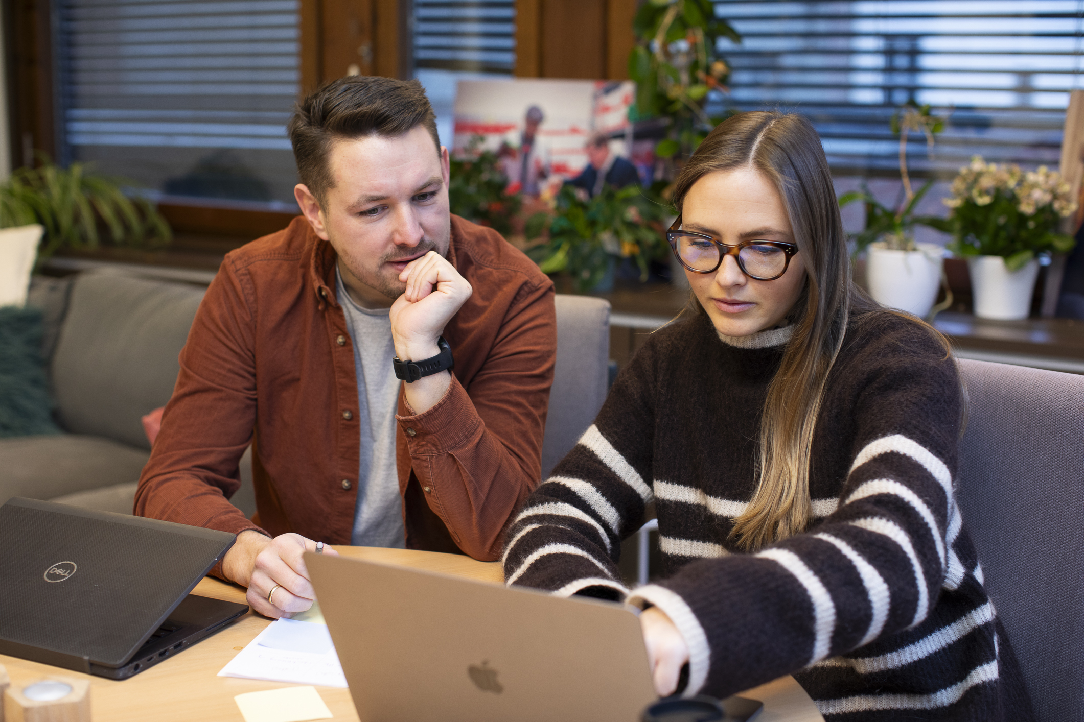 En ung mann og en kvinne sitter ved et bord og jobber på hver sin PV. De har begge briller. Hun har langt, mørkt hår og stripete genser