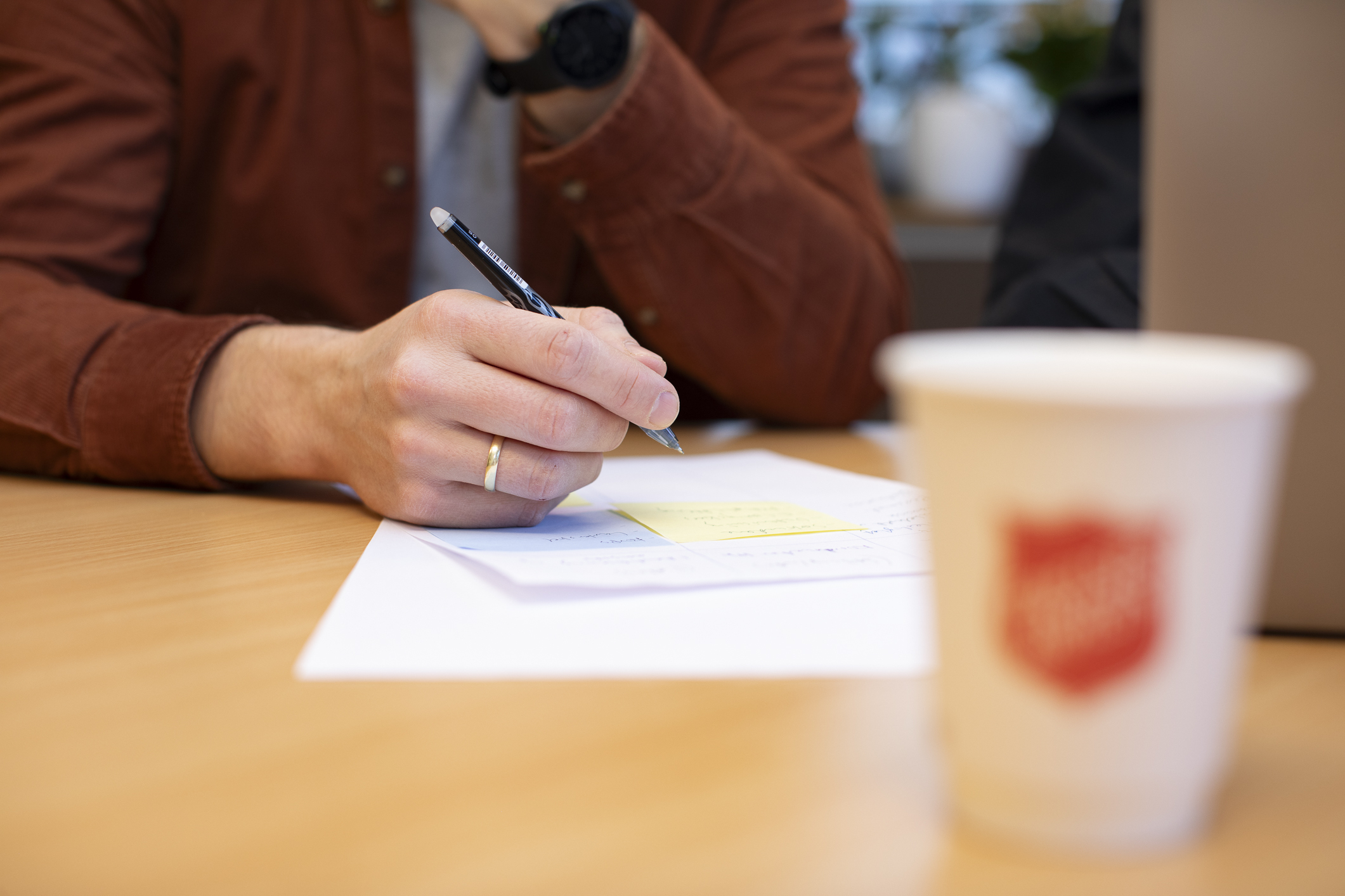 En mannlig hånd skriver på et ark på et bord. En pappkopp med Frelsesarmeen på er i forgrunnen på bildet.
