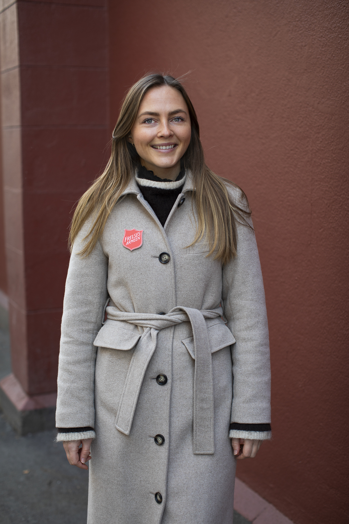 Ung kvinne med langt, mørkt hår og briller med en grå ullkåpe og en rød vegg i bakgrunnen.