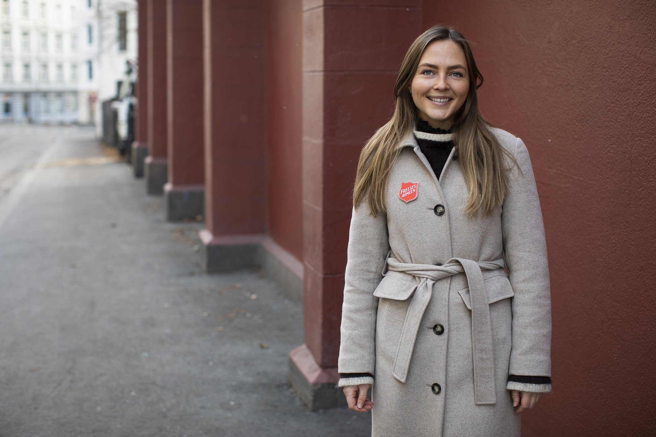 Ung kvinne med langt, mørkt hår og briller med en grå ullkåpe og en rød vegg i bakgrunnen.
