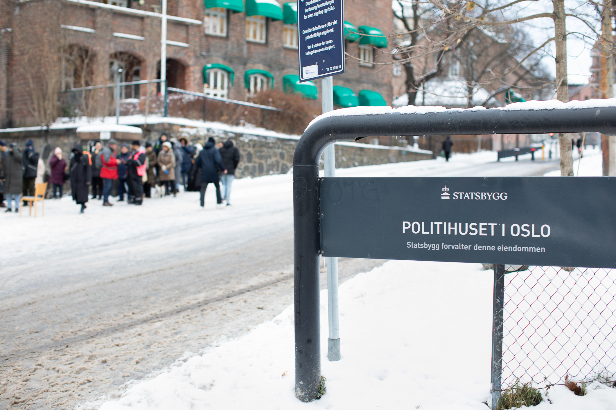 Et grønt skilt i forgrunnen  der det står Politihuset i Oslo. Det er vinter og mye snø i gaten I bakgrunnen ser man en rekke folk med boblejakker som står i kø.