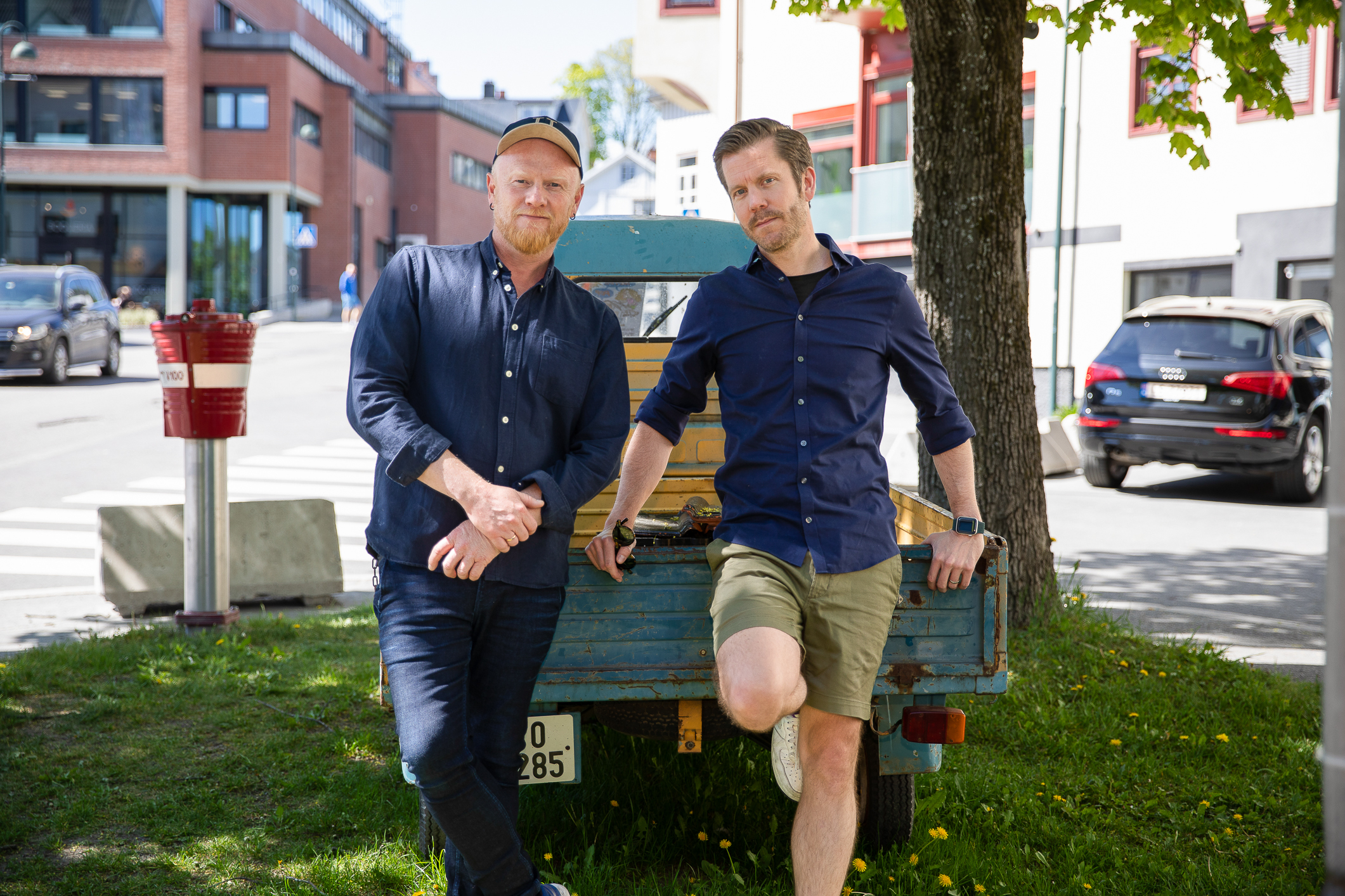 To menn står lent opp mot en bil ute i en liten by.