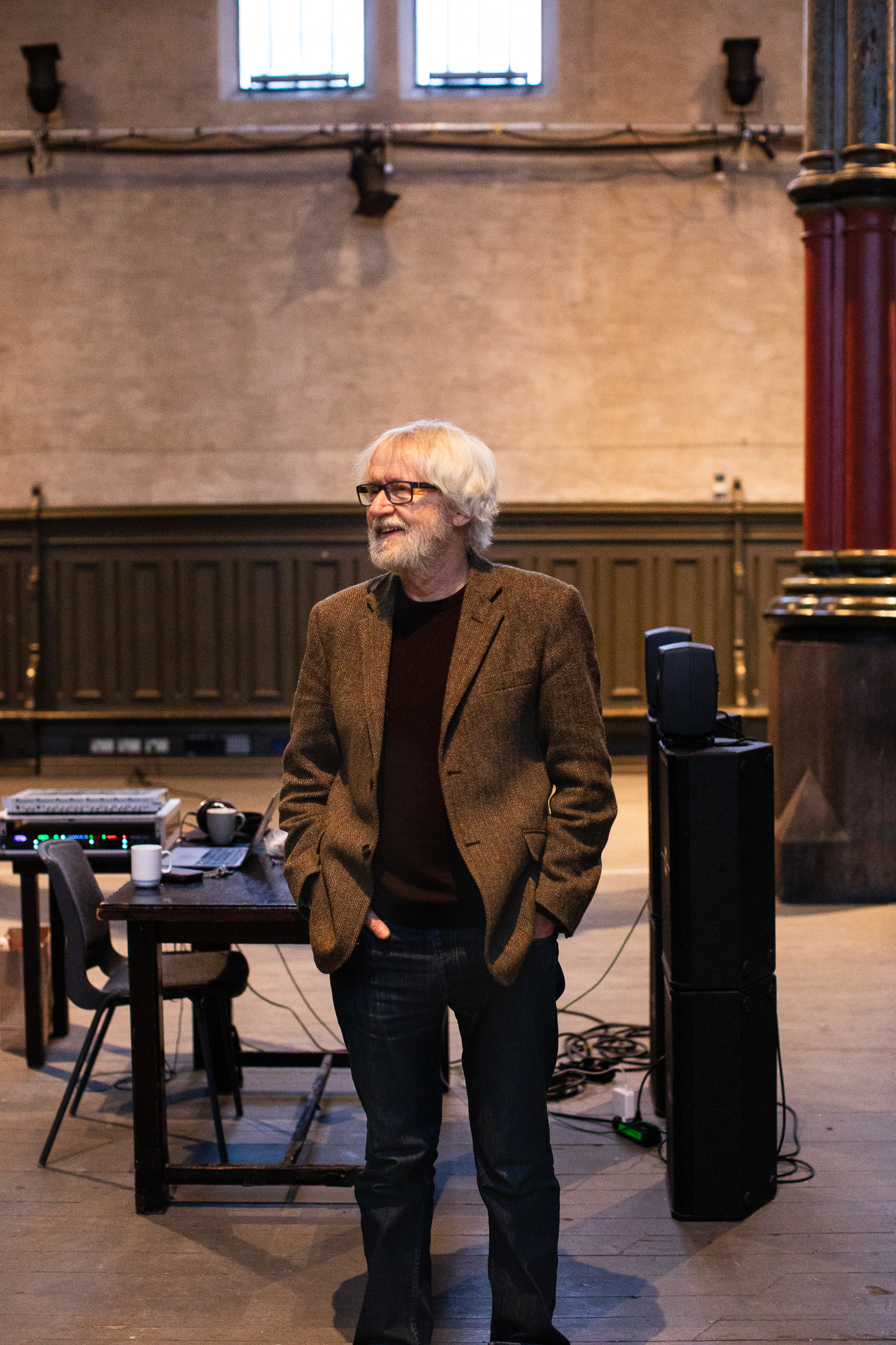 Gråhåret mann med brun tweedjakke står alene i et stort kirkerom i brune toner.